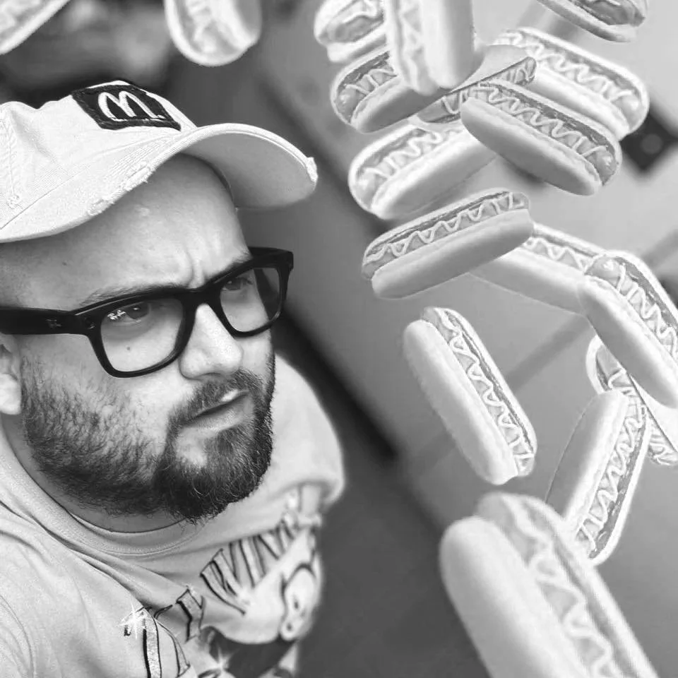A man with a beard, glasses, and a McDonald's hat looking at a floating row of hot dogs with mustard and relish.
