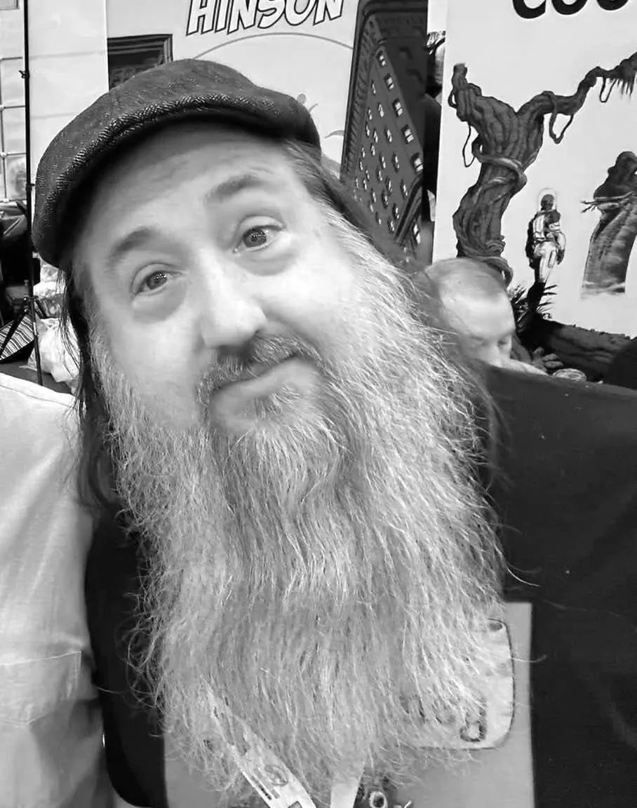 A man with a long beard and mustache wearing a flat cap, looking slightly upward in a black and white photo, with artwork and signs in the background.