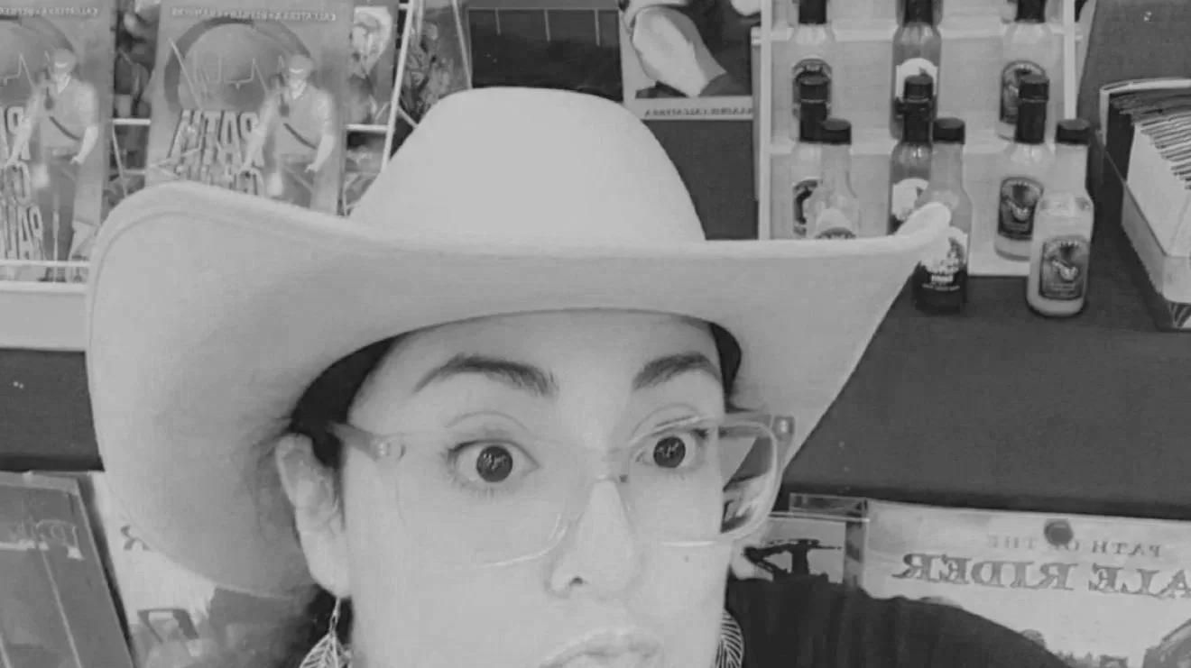 A woman wearing a cowboy hat and glasses taking a selfie in a store aisle with various health and wellness products in the background.