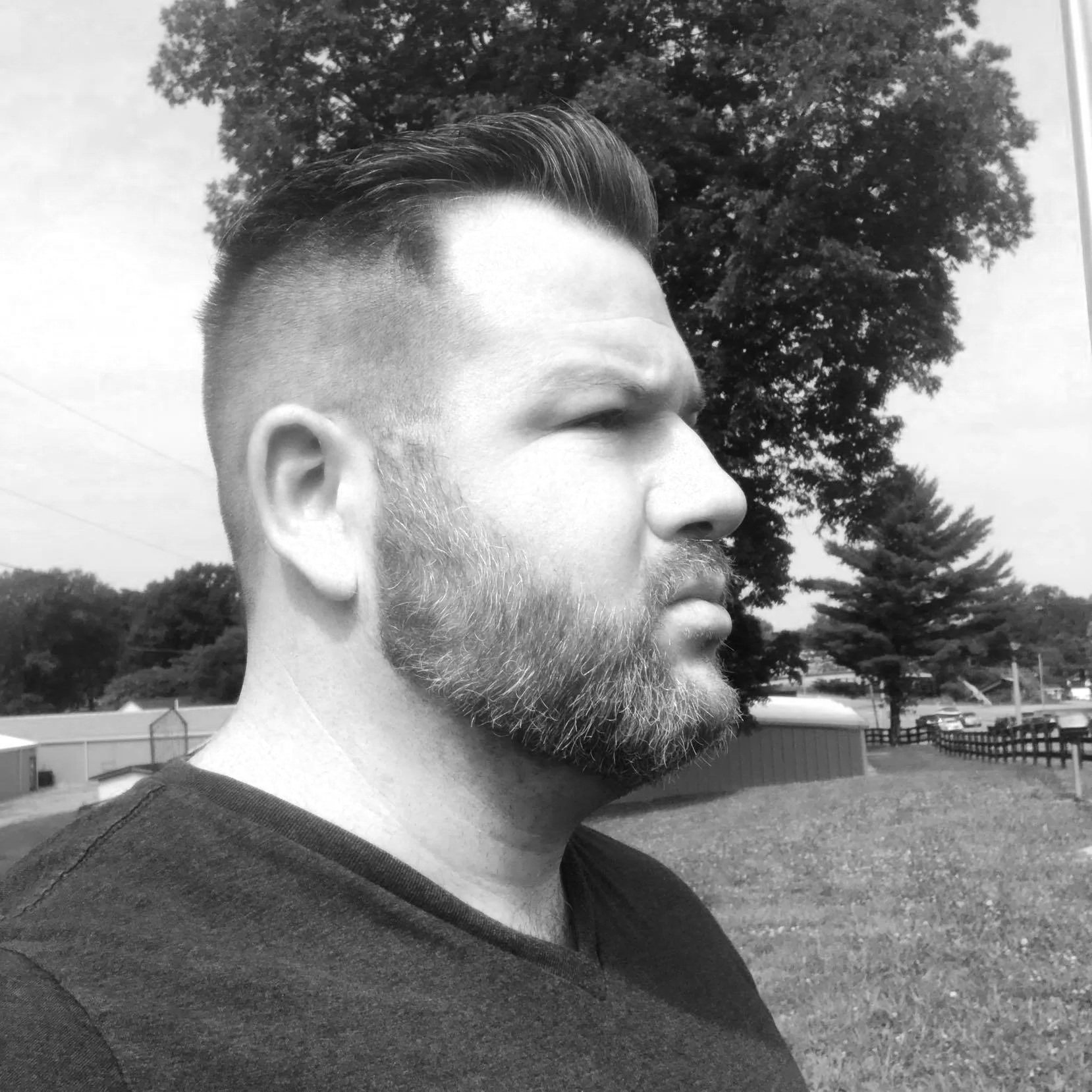 Side profile of a bearded man with styled hair standing outdoors in a grassy area with trees and fences in the background, in black and white.