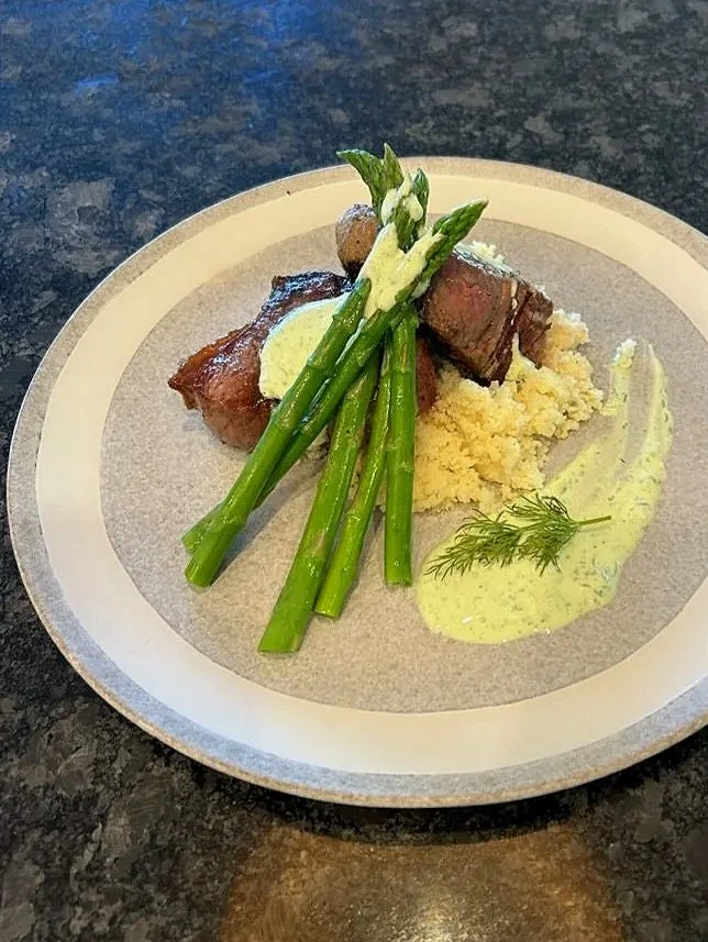 Plate with grilled steak topped with herb butter, cooked asparagus spears, mashed potatoes with herb sauce, and a sprig of dill on a light-colored plate.