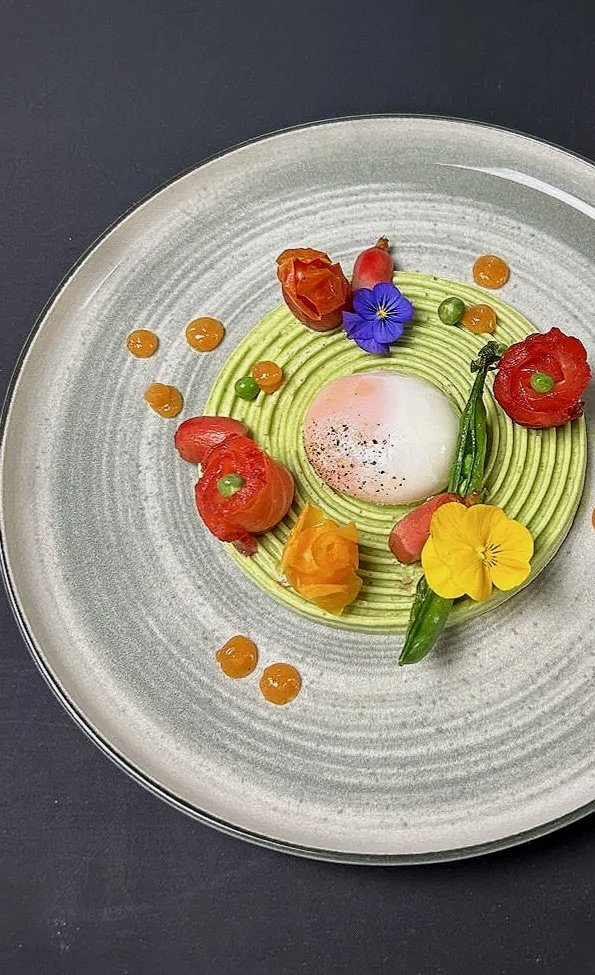 A gourmet dish with a poached egg at the center, topped with black pepper, surrounded by green puree, edible flowers, small tomatoes, and green peas on a gray plate.