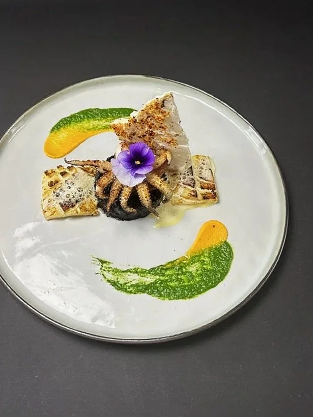 A plated gourmet dish featuring a piece of fish or seafood garnished with an edible purple flower, served with colorful sauce streaks in green, yellow, and orange, on a white round plate.