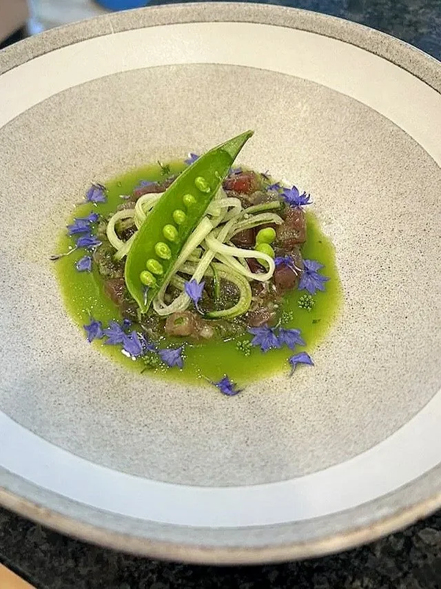 A gourmet dish with thinly sliced raw fish, likely tuna or another sashimi, topped with a sugar snap pea, green sauce, purple edible flowers, and garnished with thinly sliced vegetables on a textured beige plate.