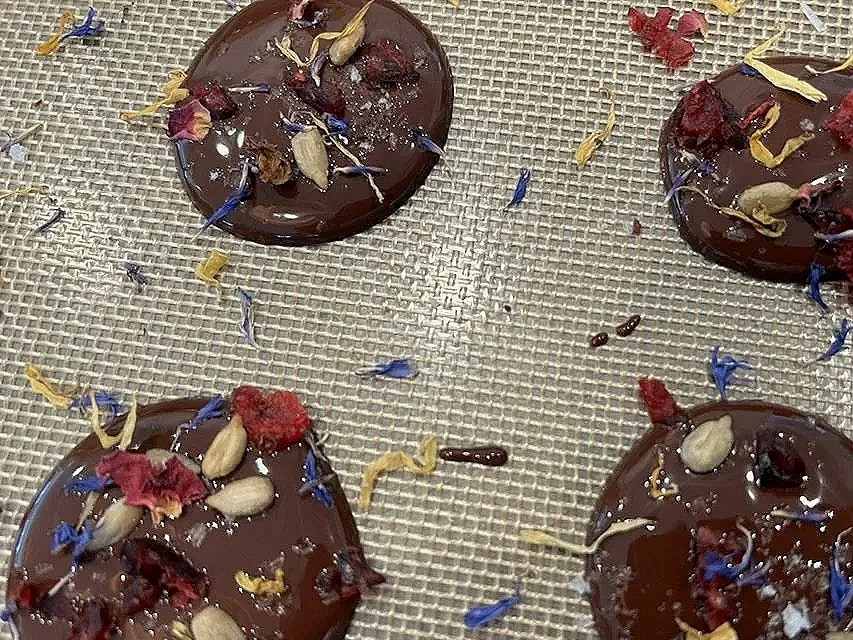 Four round chocolates topped with dried flowers and nuts, placed on a golden mesh surface.