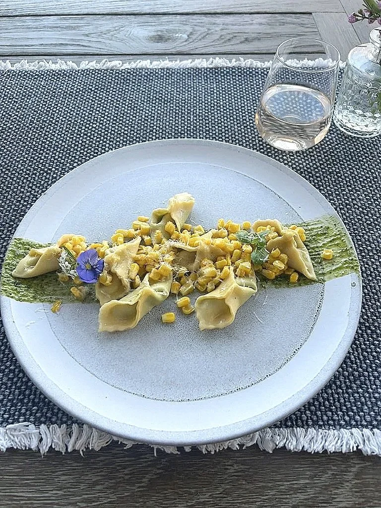 Plate of pasta with ravioli topped with corn, garnished with edible flowers and green sauce on a white plate, with a glass of water and a small vase of flowers in the background.