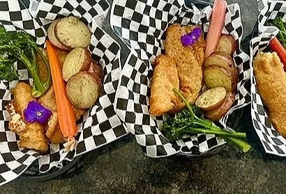 Three fish tacos with fried fish fillets, tomatoes, lettuce, and purple edible flowers, served with roasted potatoes and fresh vegetables in checkered baskets.