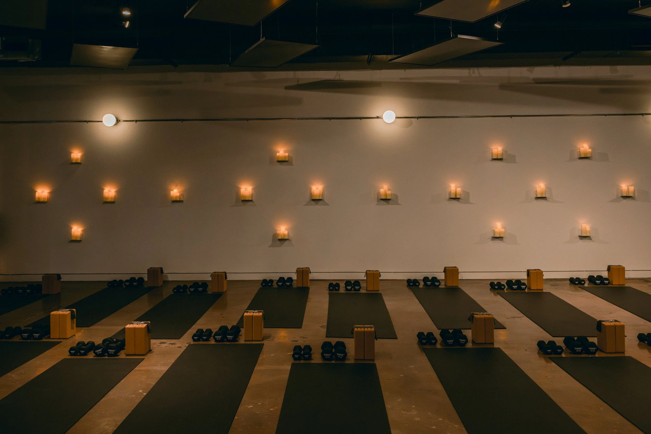 A yoga and pilates studio, with black mats on the floor, candlelight, and small black and blue dumbbells placed on racks. The wall features many small wall-mounted lights, creating a warm ambiance.