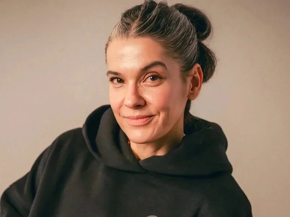 A female founder of yoga studio with gray hair tied back, wearing a black hoodie, smiling at the camera against a plain neutral background.