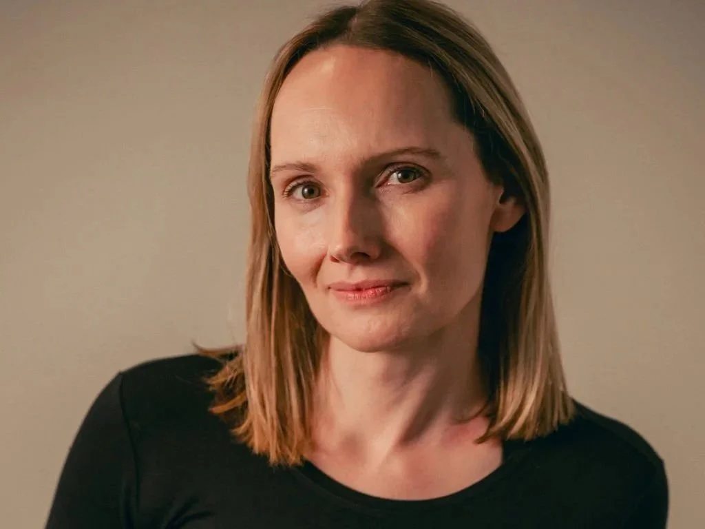 A female founder of pilates studio with shoulder-length blonde hair and a slight smile, wearing a black top, standing against a plain beige background.