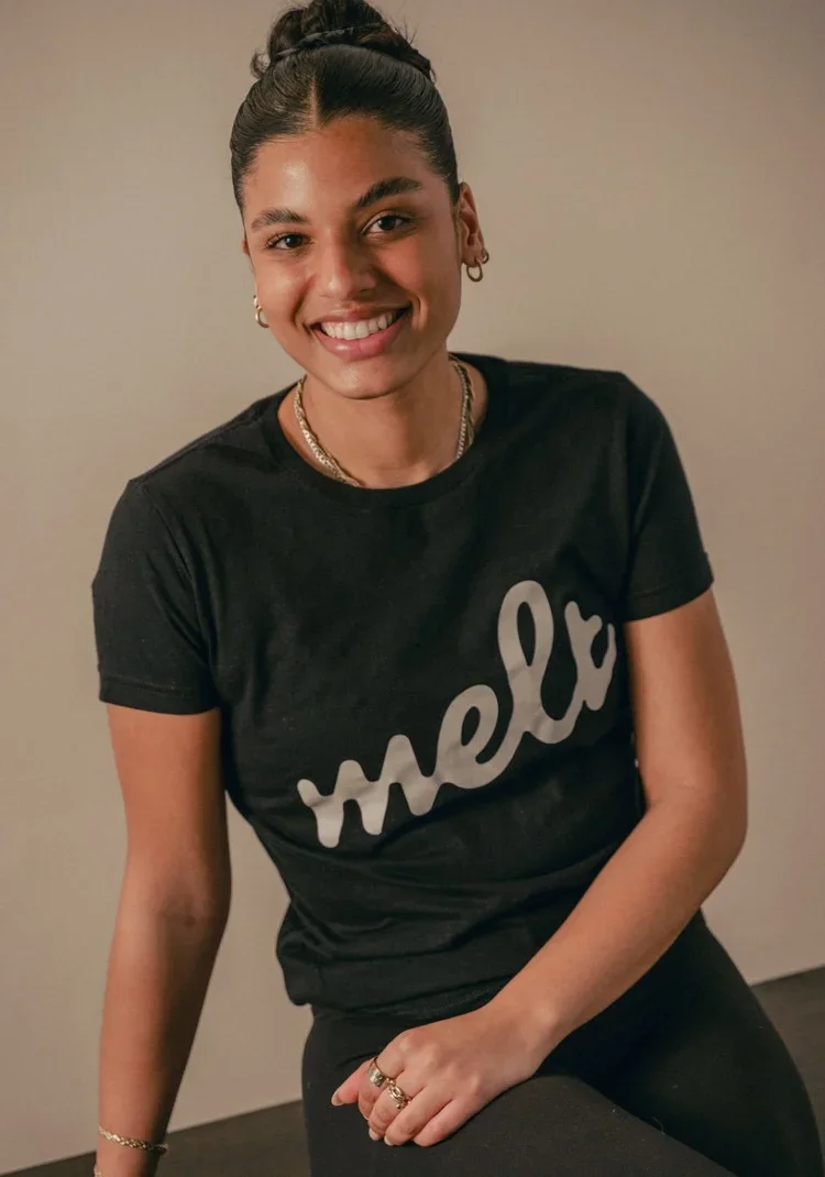 A young woman with dark hair tied up, wearing hoop earrings, layered necklaces, a black T-shirt with the word 'mel' printed on it, smiling at the camera, sitting in front of a plain gray wall.