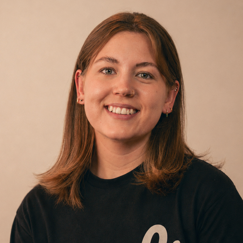 A young woman with short hair, wearing a black t-shirt with white cursive text, sitting indoors against a plain wall.