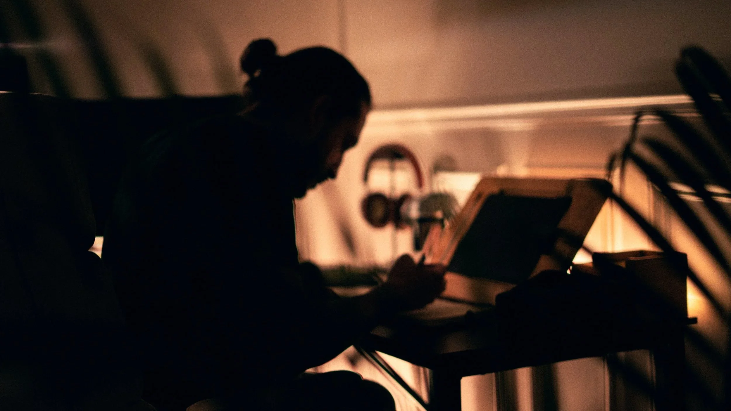 Silhouette d'une personne assise à une table, lisant un livre dans une pièce sombre, avec un éclairage tamisé chaleureux.