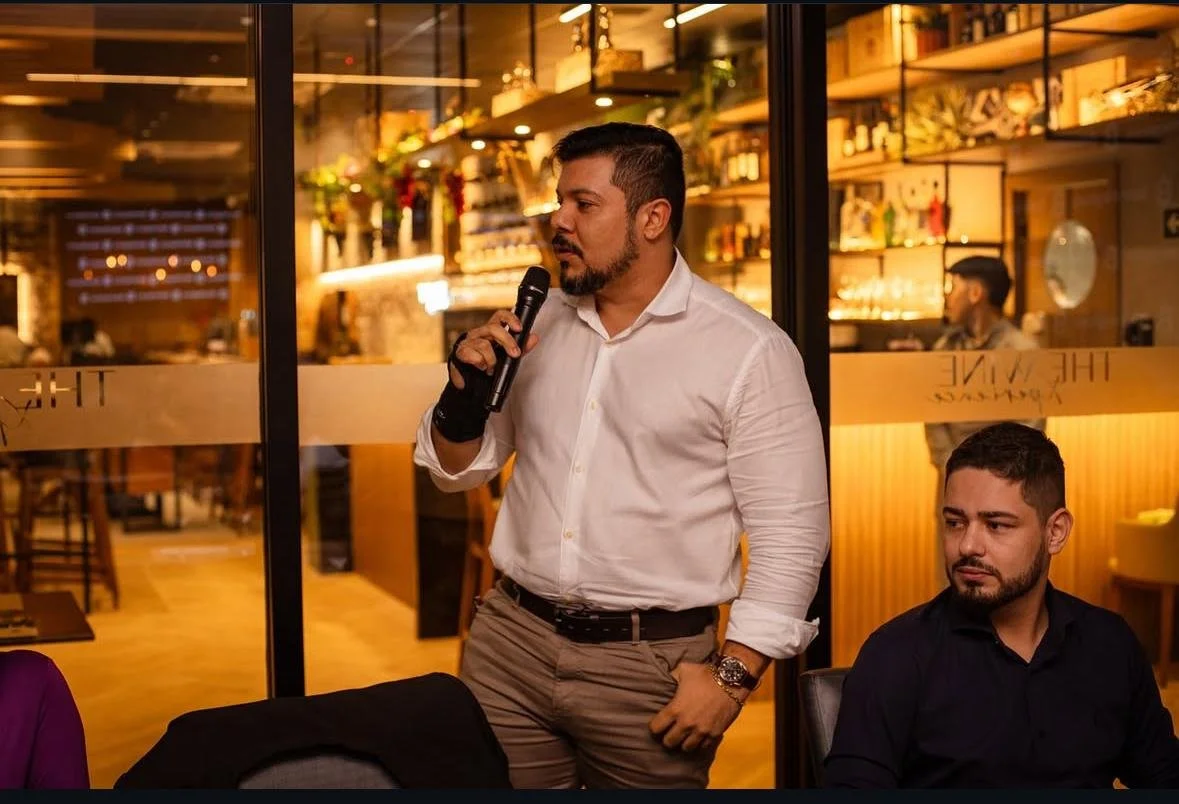 Homem com barba e cabelo curto, vestindo camisa branca de manga longa e calça bege, segurando um microfone, aparentemente falando ou apresentando algo, ao lado de outro homem de cabelo curtinho, barba por fazer, vestindo camisa preta, sentado numa mesa, em um ambiente interno de restaurante ou bar com decoração quente e iluminação amarelada.