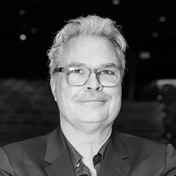 Schwarz-Weiß-Foto eines Mannes mit Brille, der in einem Anzug lächelnd in die Kamera schaut.