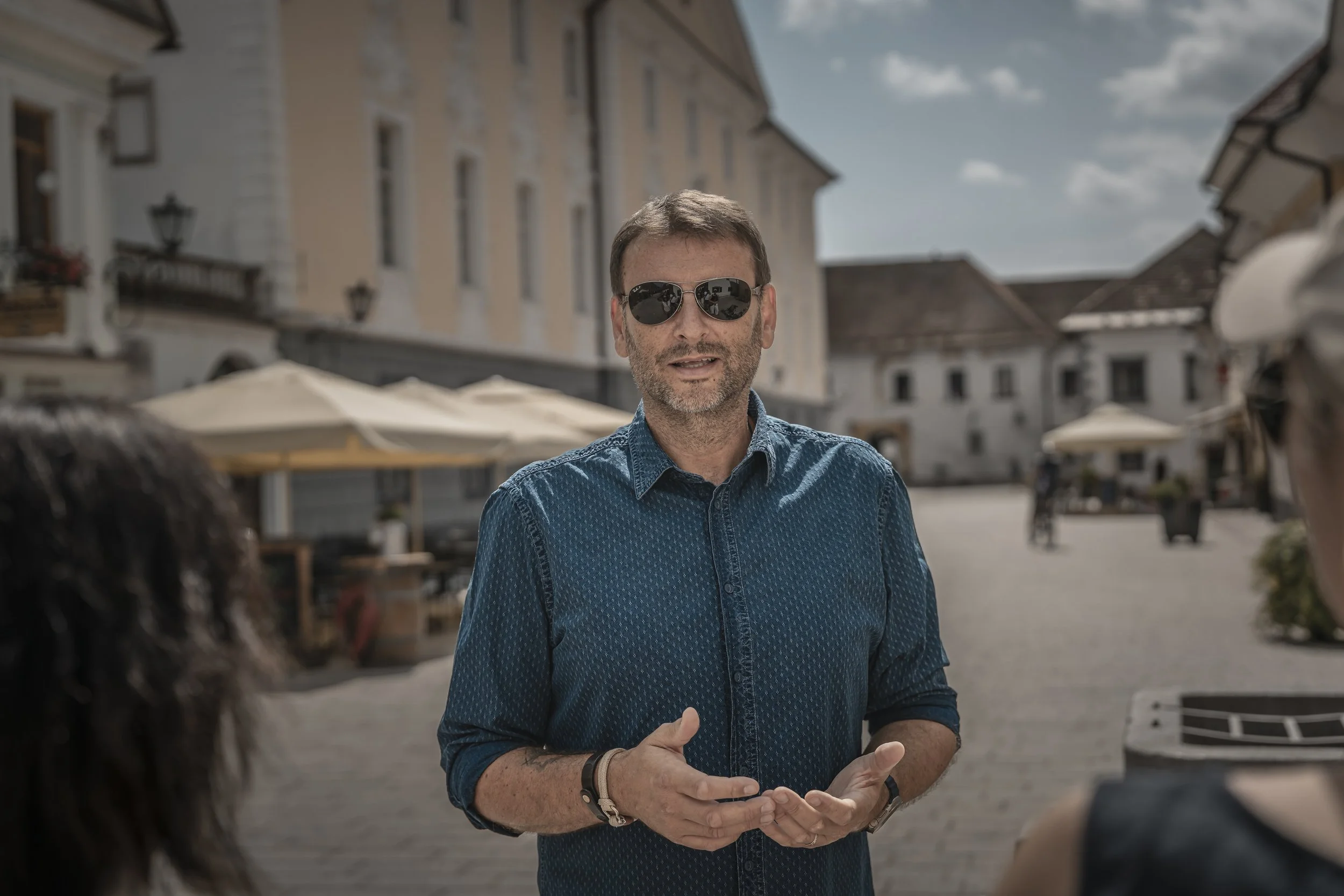 A male tour guide, speaking to a group in an outdoor setting with European-style buildings and umbrellas in the background.