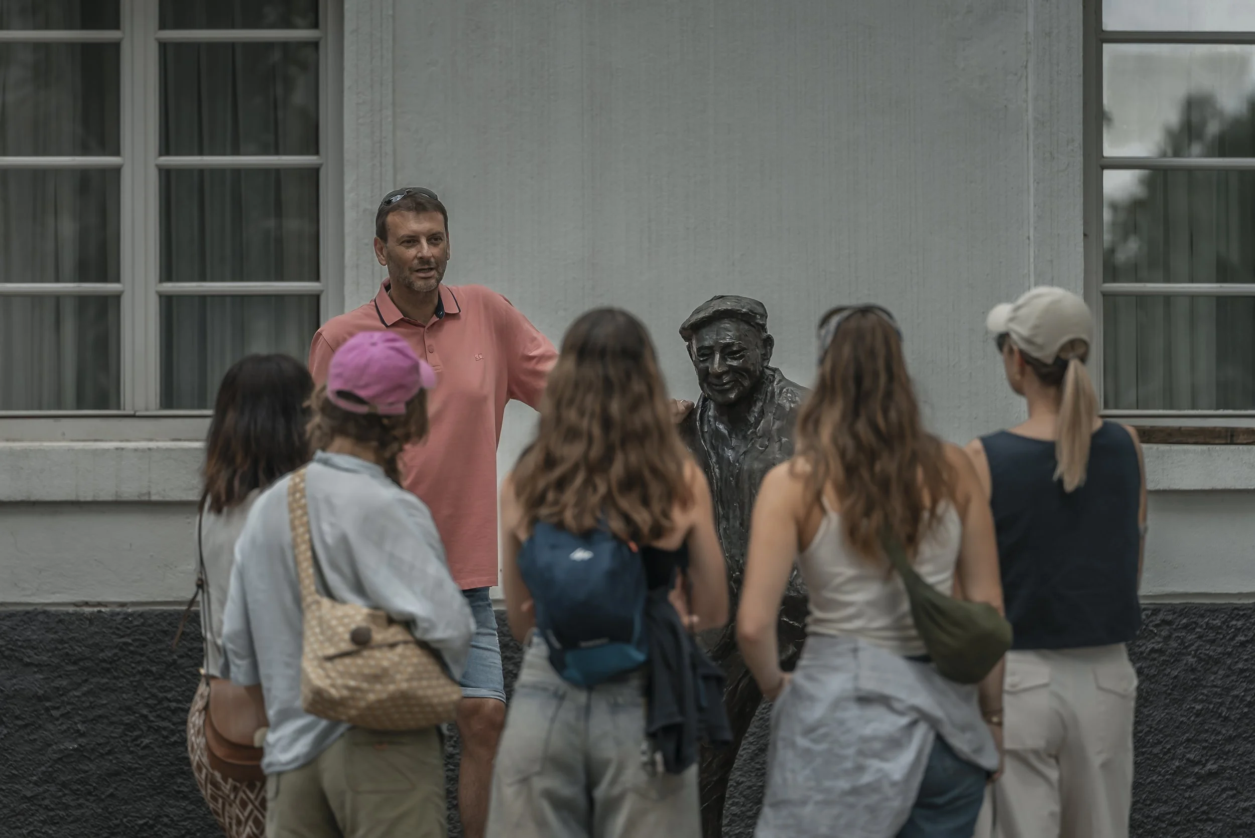 A man wearing a pink shirt is giving a tour or lecture to a group gathered around a bust statue outside a building with large windows.