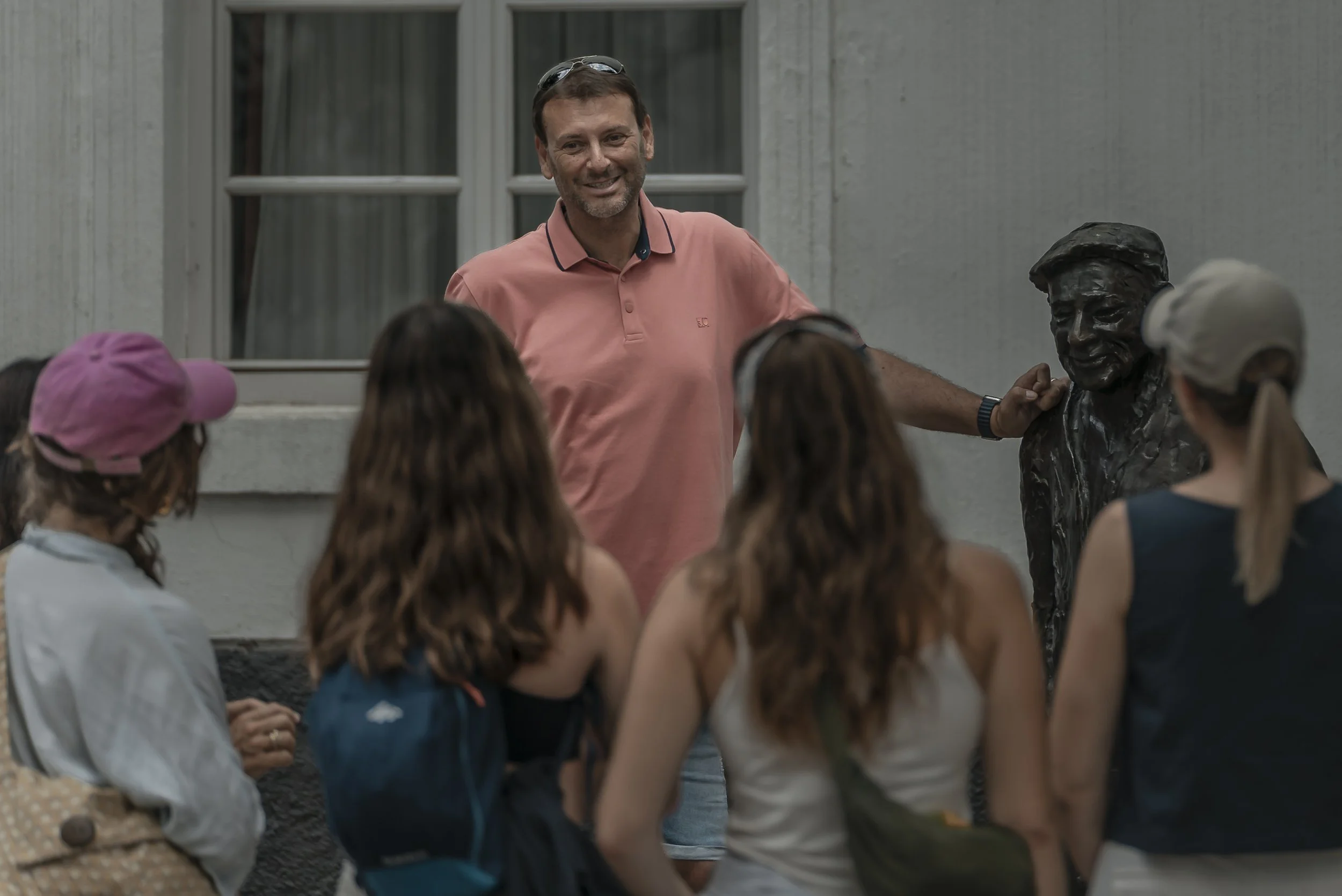 A tour guide in a pink polo shirt is smiling and standing outdoors, surrounded by a group of people, with a bronze bust sculpture beside him.