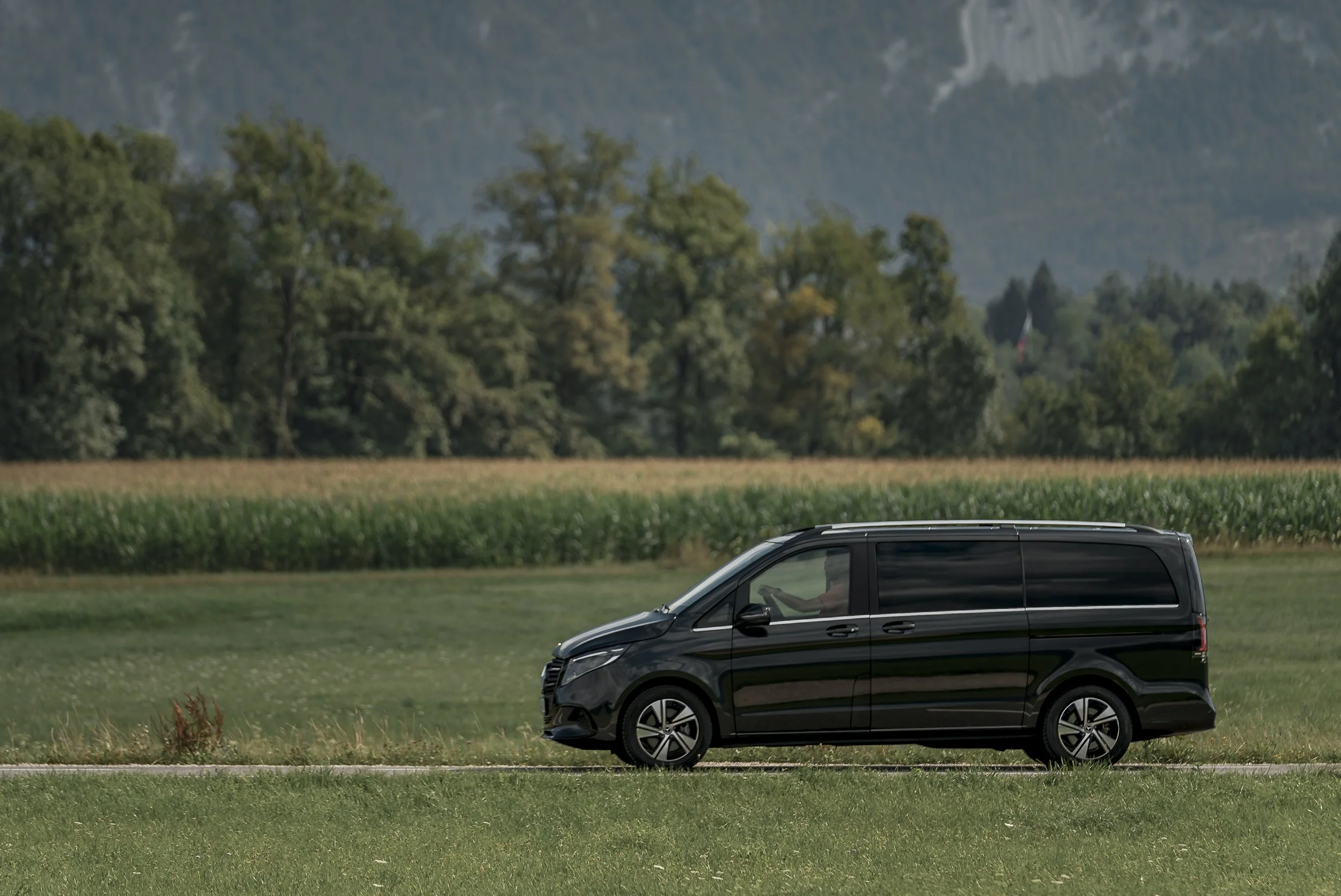 A black van driving on a rural road with green fields and trees, and mountains in the background.