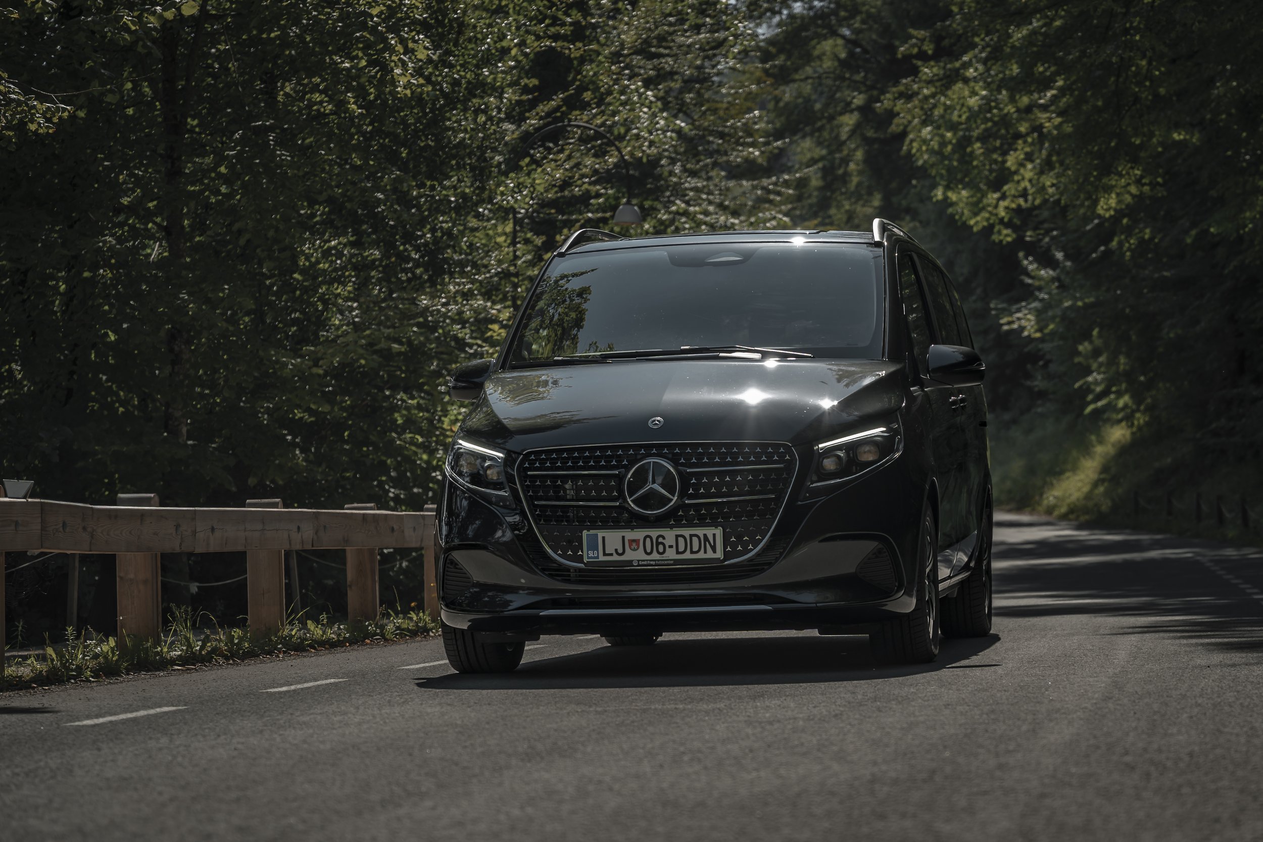 Black Mercedes-Benz van driving on a forested road with green trees and a wooden guardrail on the side.