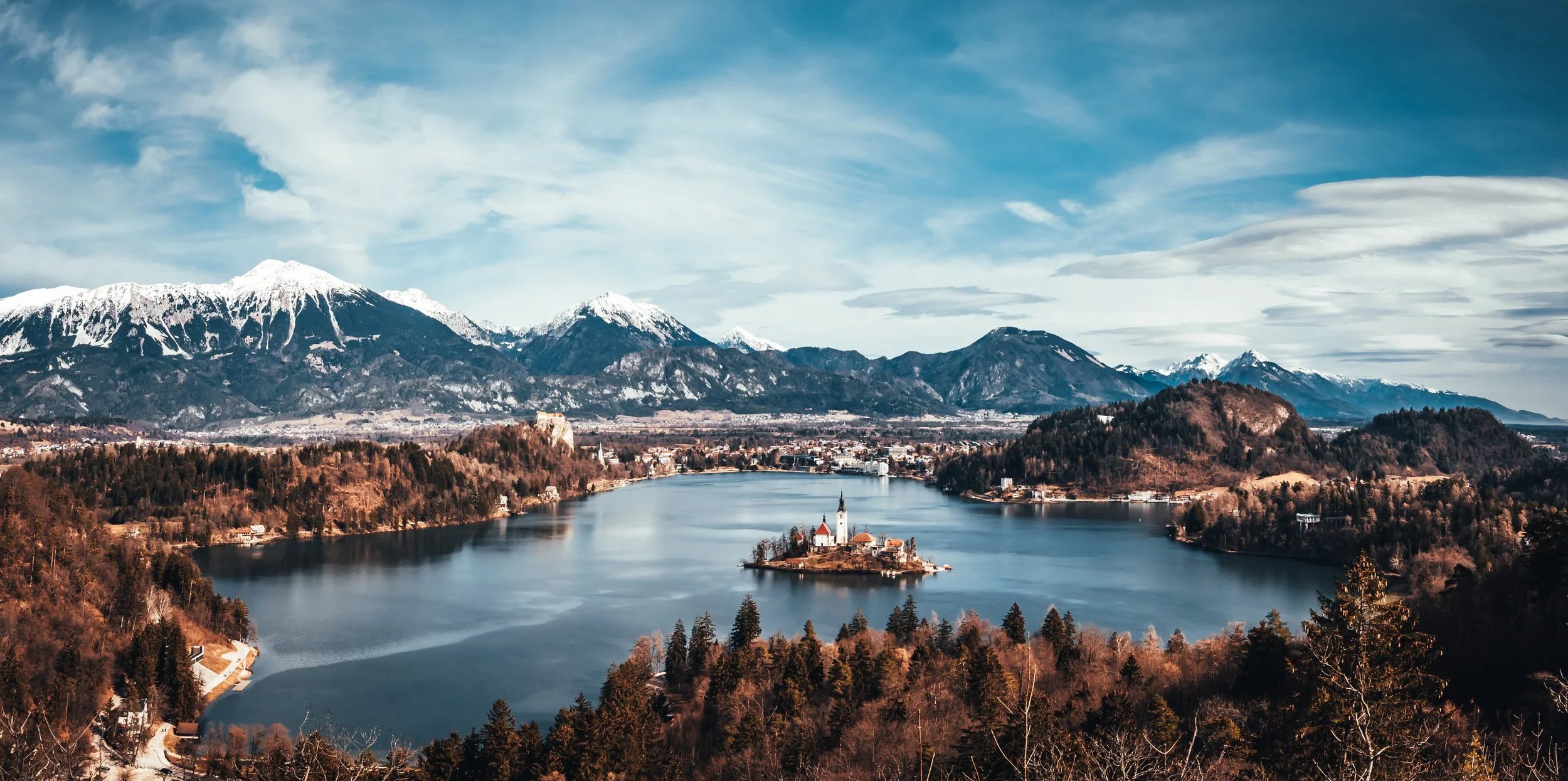A scenic view of Lake Bled, featuring an island with a church, surrounded by forest and mountains with snow-capped peaks in the background, under a partly cloudy sky.