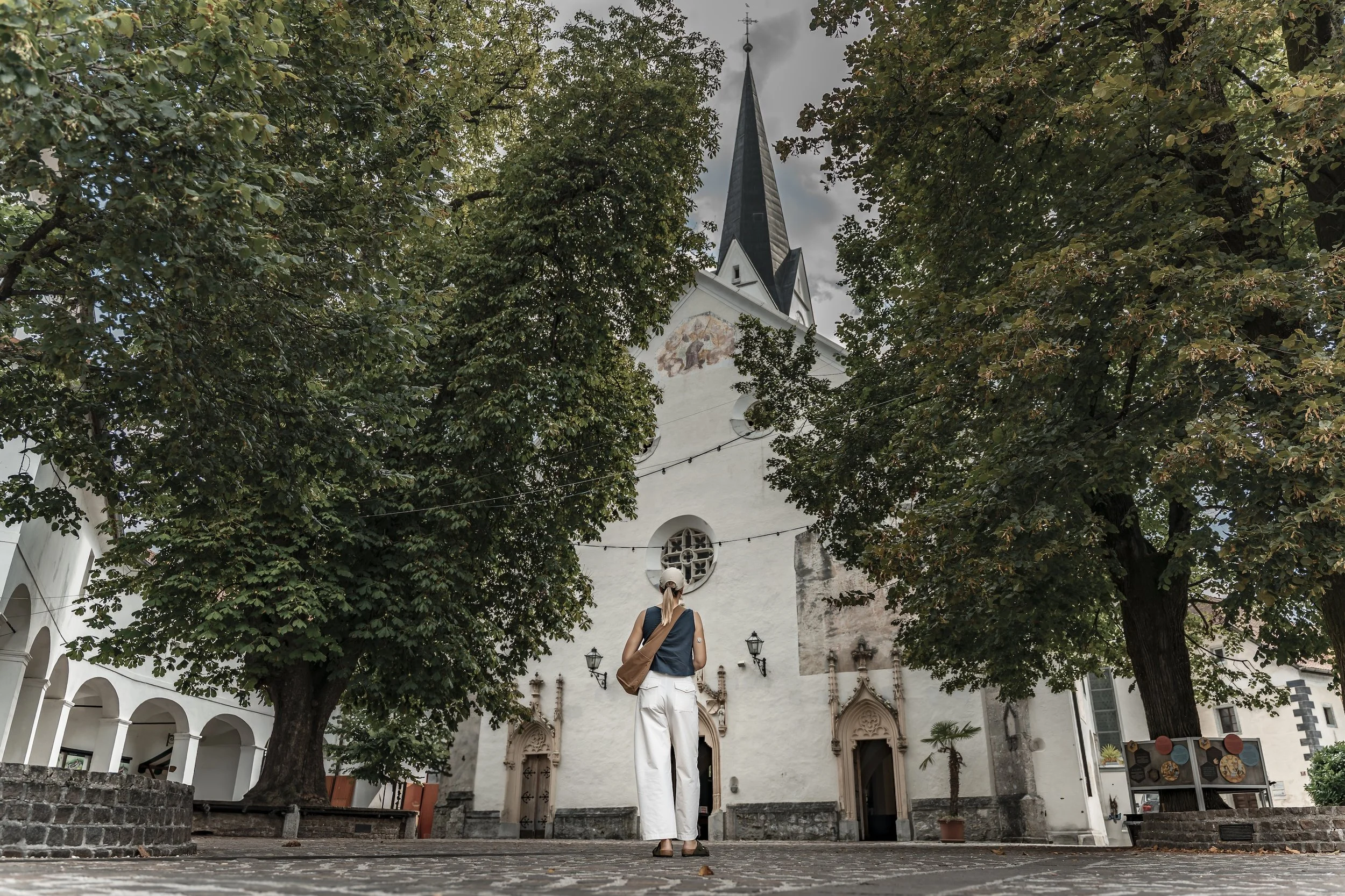 A woman standing in front of a historic white church with a tall steeple, surrounded by large trees and cobblestone pavement.