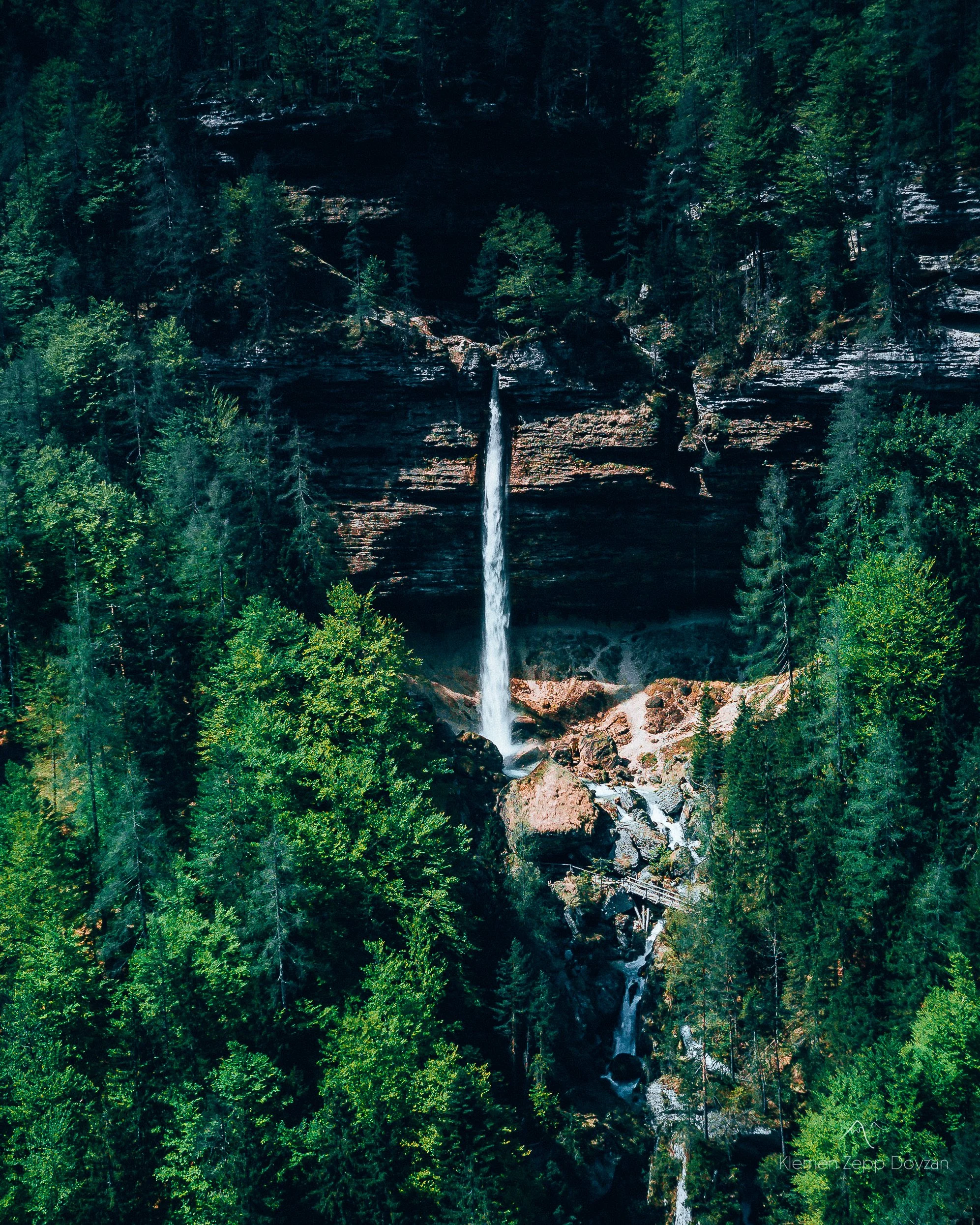 Tall waterfall cascading down a rocky cliff surrounded by dense evergreen forest.