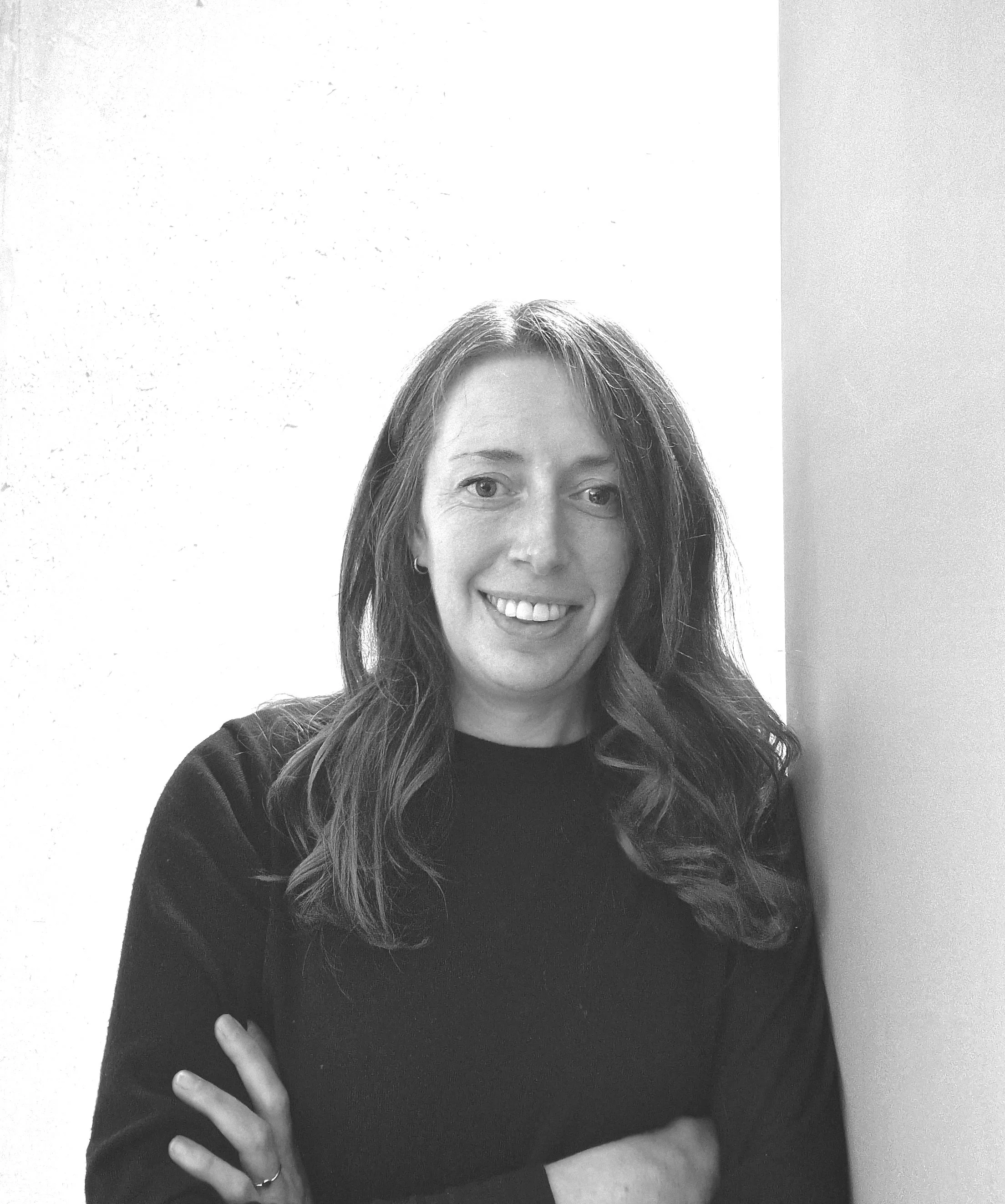A black and white photo of a woman with long wavy hair, smiling, with her arms crossed, standing against a plain light-colored wall.