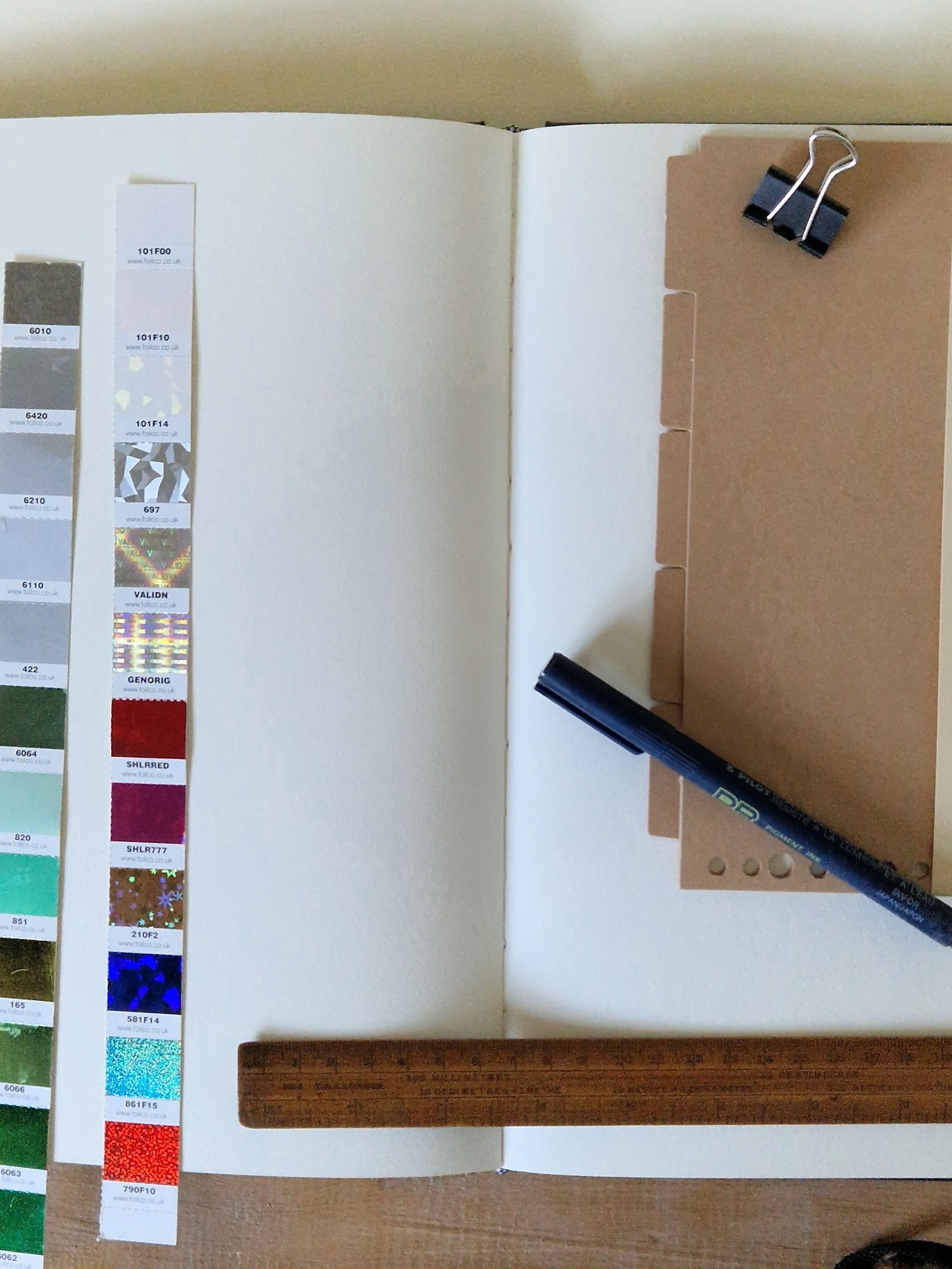 Open scrapbook with color swatches on the left and a brown wooden sample on the right, secured by black binder clips and a black marker.