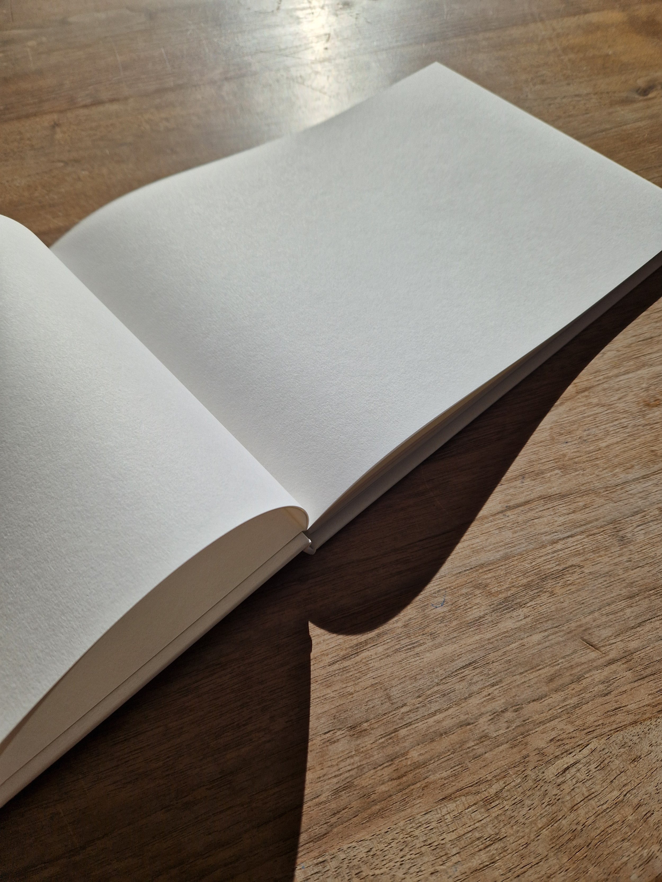 Open blank white notebook on a wooden surface with sunlight and shadows.