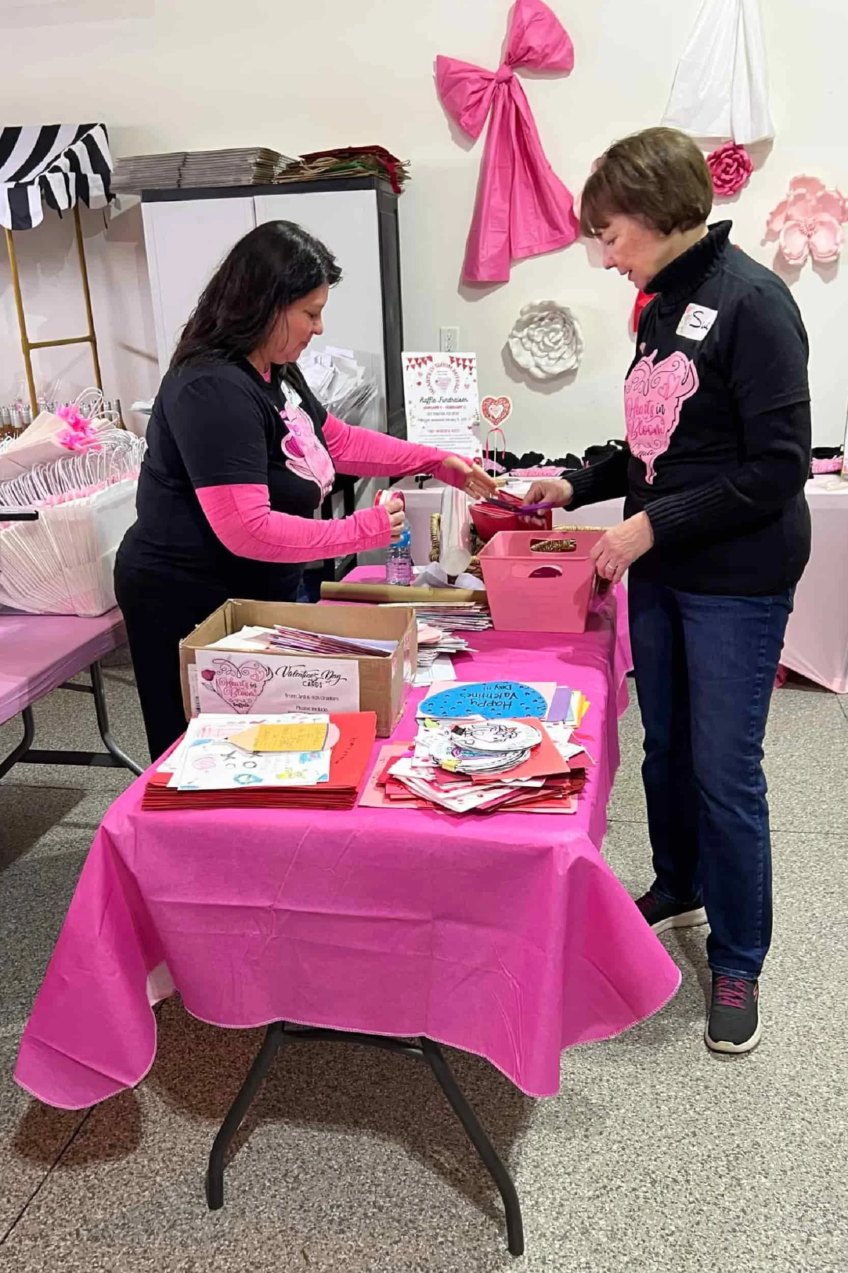 Two women at a pink-themed Valentine's Day event table, organizing and selling Valentine's Day cards and related items.
