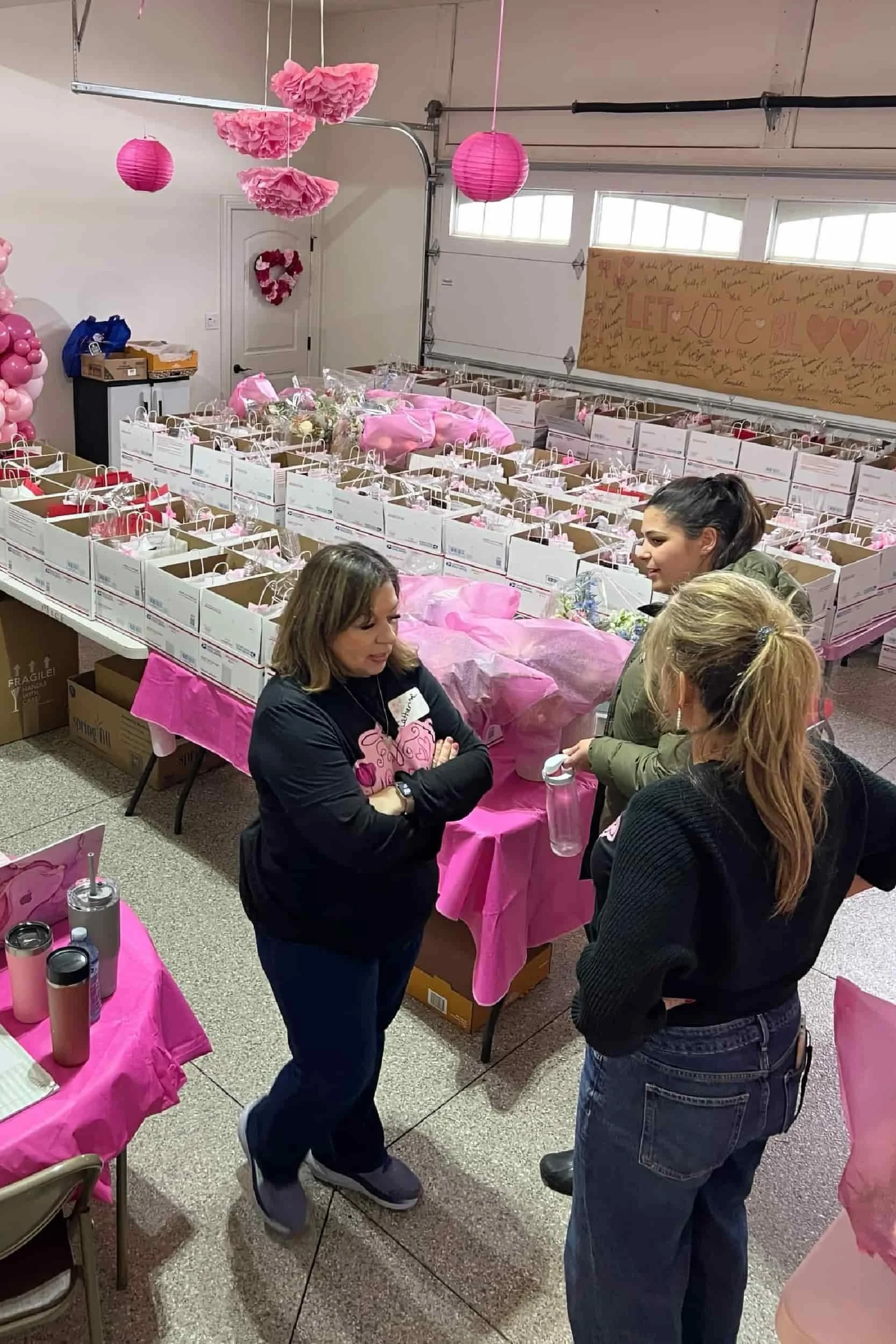 Three women stand in a decorated room with pink theme, surrounded by boxes filled with pink items and pink decorations hanging from the ceiling, preparing for an event.