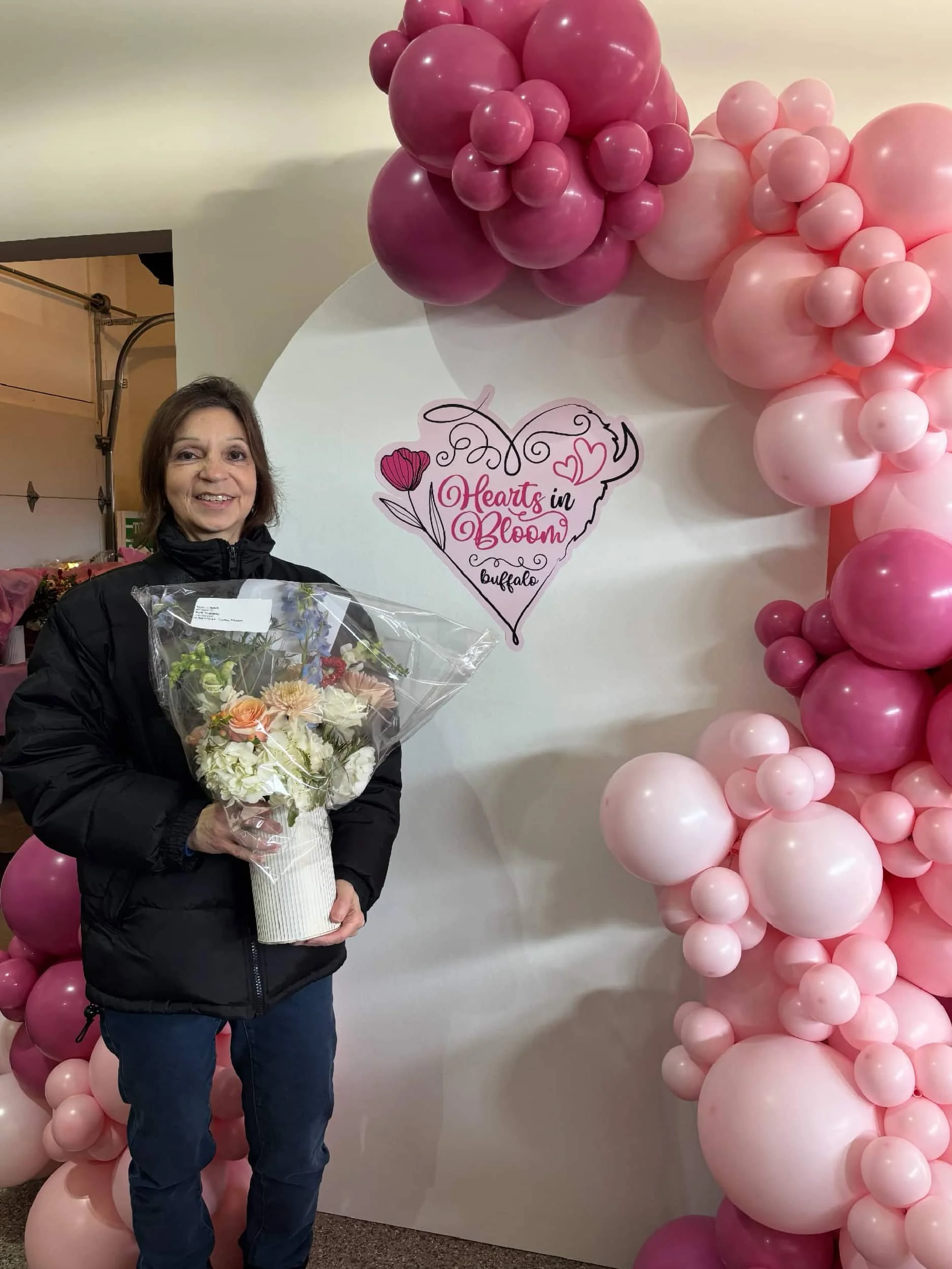 Woman holding a bouquet of flowers standing in front of a backdrop decorated with pink and purple balloons for Hearts in Bloom event in Buffalo.