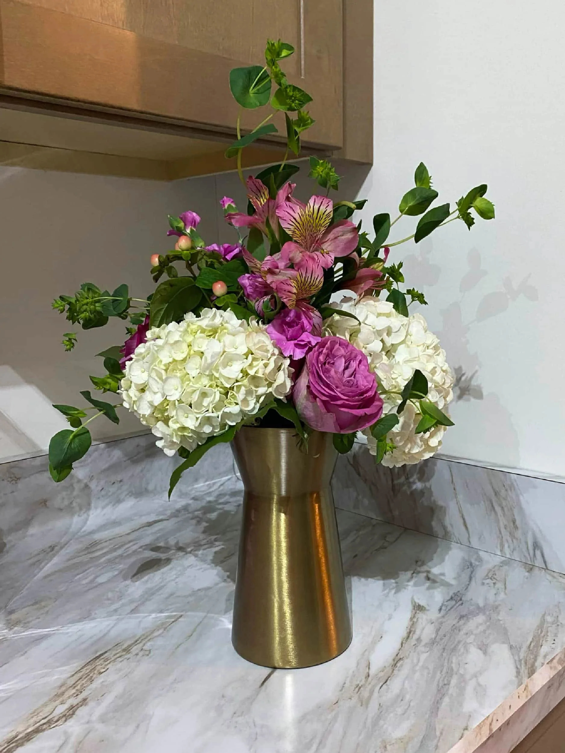 A floral arrangement with white hydrangeas, pink roses, and pink and purple lilies in a gold vase placed on a marble countertop.