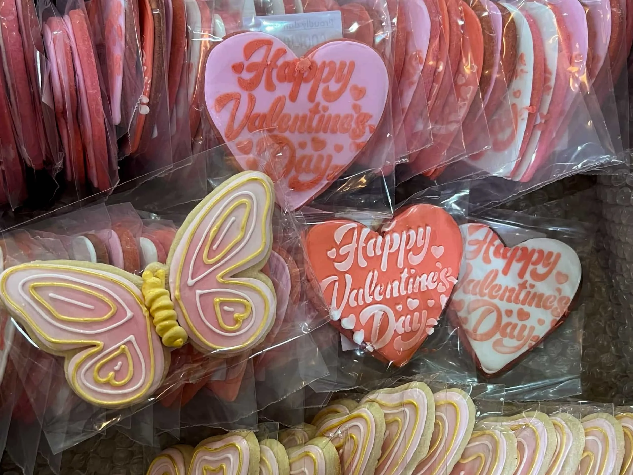 Valentine's Day cookies, including pink heart-shaped cookies with 'Happy Valentine's Day' written on them and butterfly-shaped cookies decorated with pink, white, and yellow icing, all wrapped in clear plastic