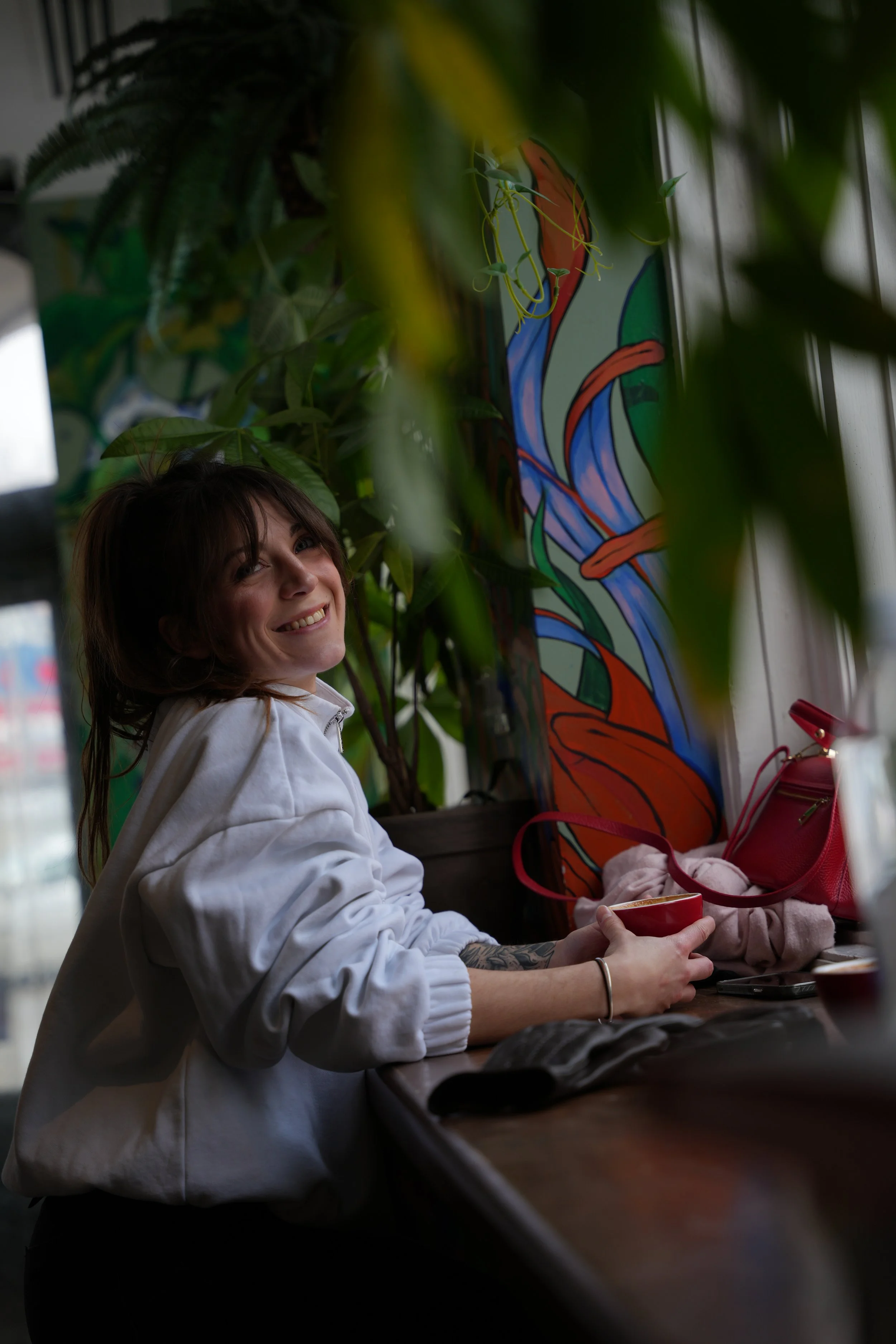 Une femme souriante avec des cheveux bruns assise à une table, entourée de plantes vertes, dans un intérieur décoré avec une peinture colorée. Elle tient une tasse rouge dans une main, en regardant vers l'objectif.