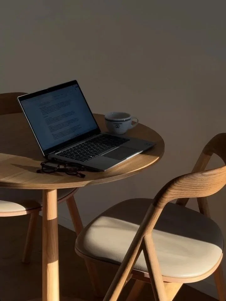 Un bureau en bois avec un ordinateur portable, une tasse et une paire de lunettes sur la table, entouré de deux chaises en bois avec des coussins beige.