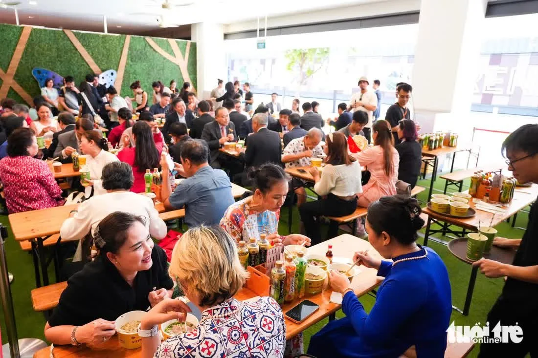 People dining together at a crowded indoor restaurant, eating food from bowls, with some drinking beverages. The restaurant has a modern interior with bright lighting and a green wall decoration.