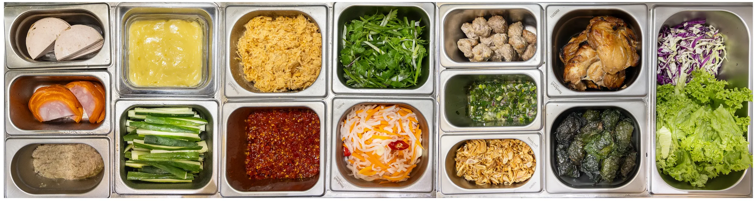 A stainless steel food counter with various compartments holding ingredients including sliced sausages, yellow pudding, shredded crab meat, fresh cilantro, chopped meat, grilled chicken, shredded cabbage and purple cabbage, sliced cucumbers, red chil