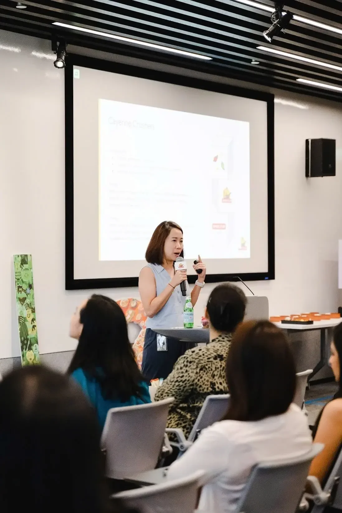 A woman giving a presentation in front of a screen while holding a microphone and standing behind a small table in a conference room with seated attendees.