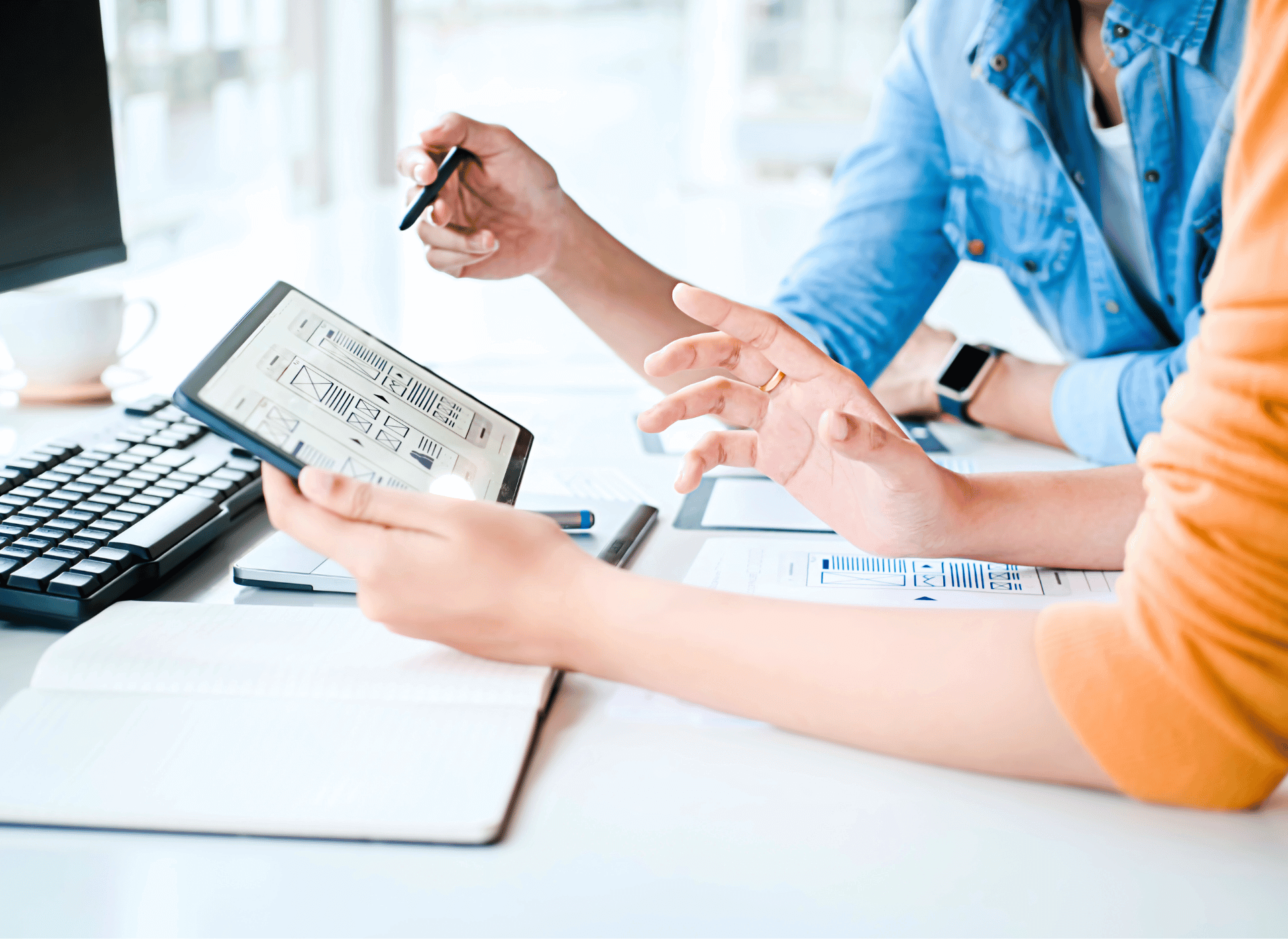 Two people discussing work at a desk, with one holding a tablet displaying a wireframe design, a keyboard, notebook, and papers.