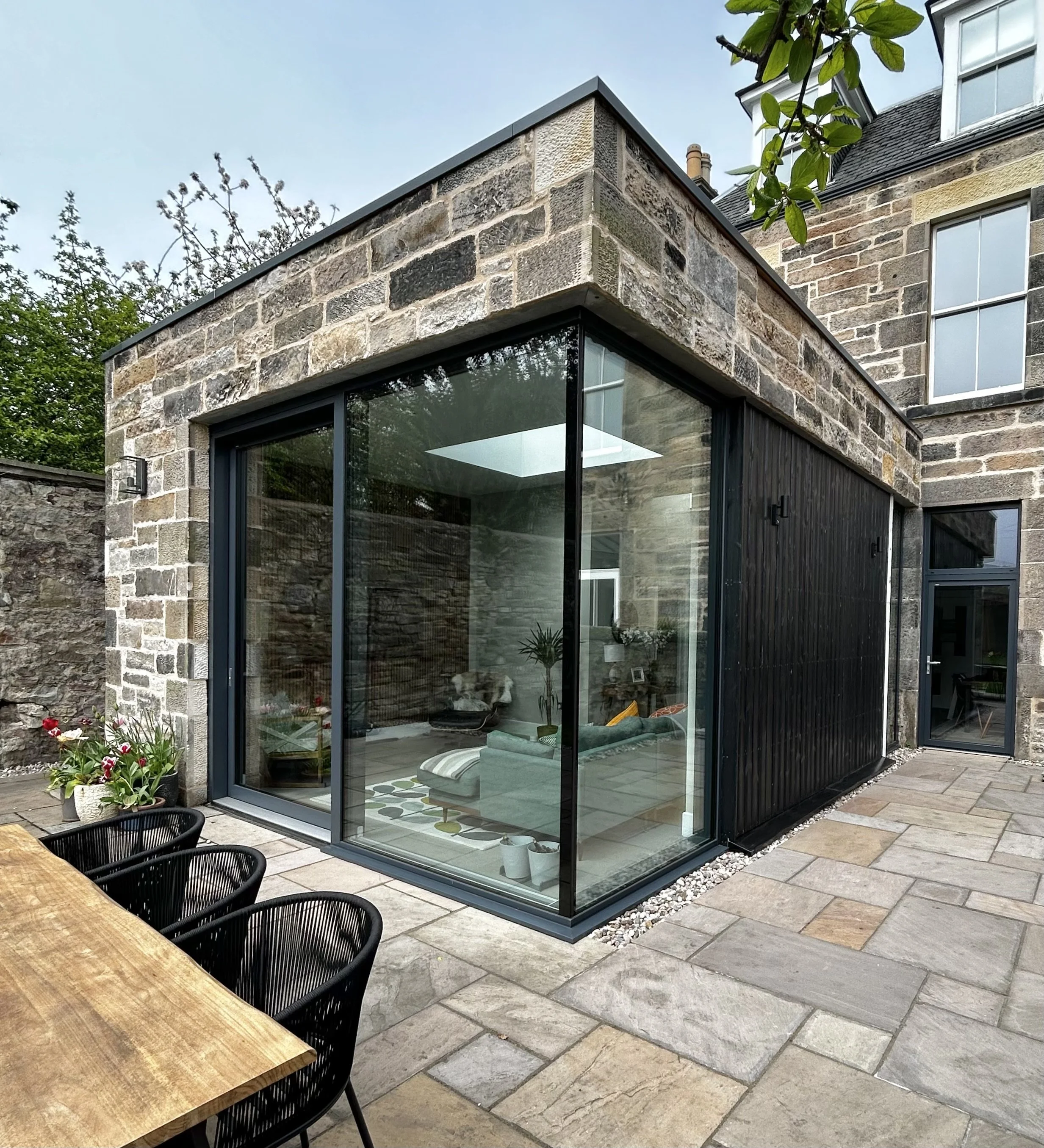 Modern extension with glass walls on a stone house, overlooking a patio with outdoor chairs and a wooden table, surrounded by a stone wall and courtyard plants.