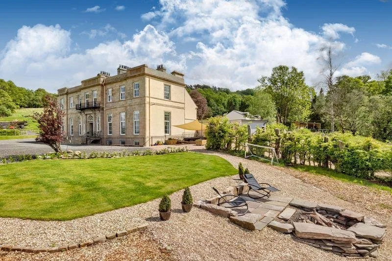 Large historic beige mansion with a landscaped backyard featuring a grassy area, gravel pathway, potted plants, and outdoor seating with lounge chairs and a fire pit, surrounded by trees under a partly cloudy sky.