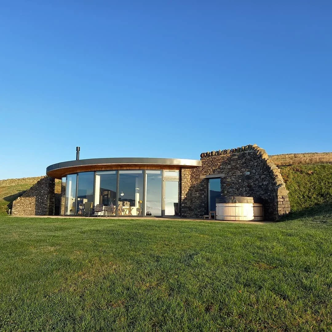 Modern house with large glass windows, stone walls, and a grassy lawn under a clear blue sky.