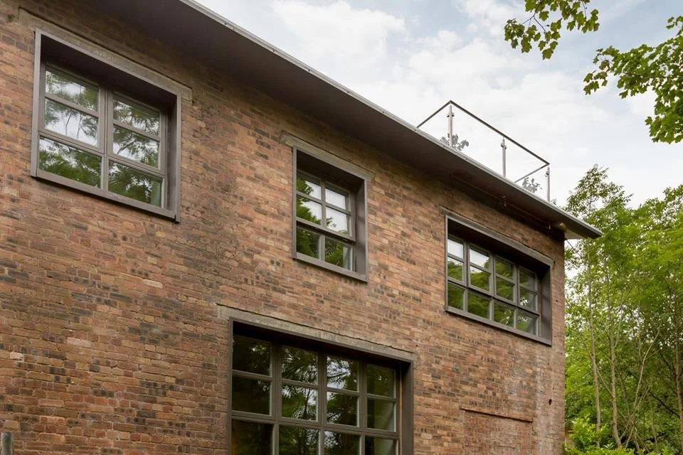 Brick residential building with four black-framed windows and a rooftop terrace surrounded by a metal railing, amidst green trees.