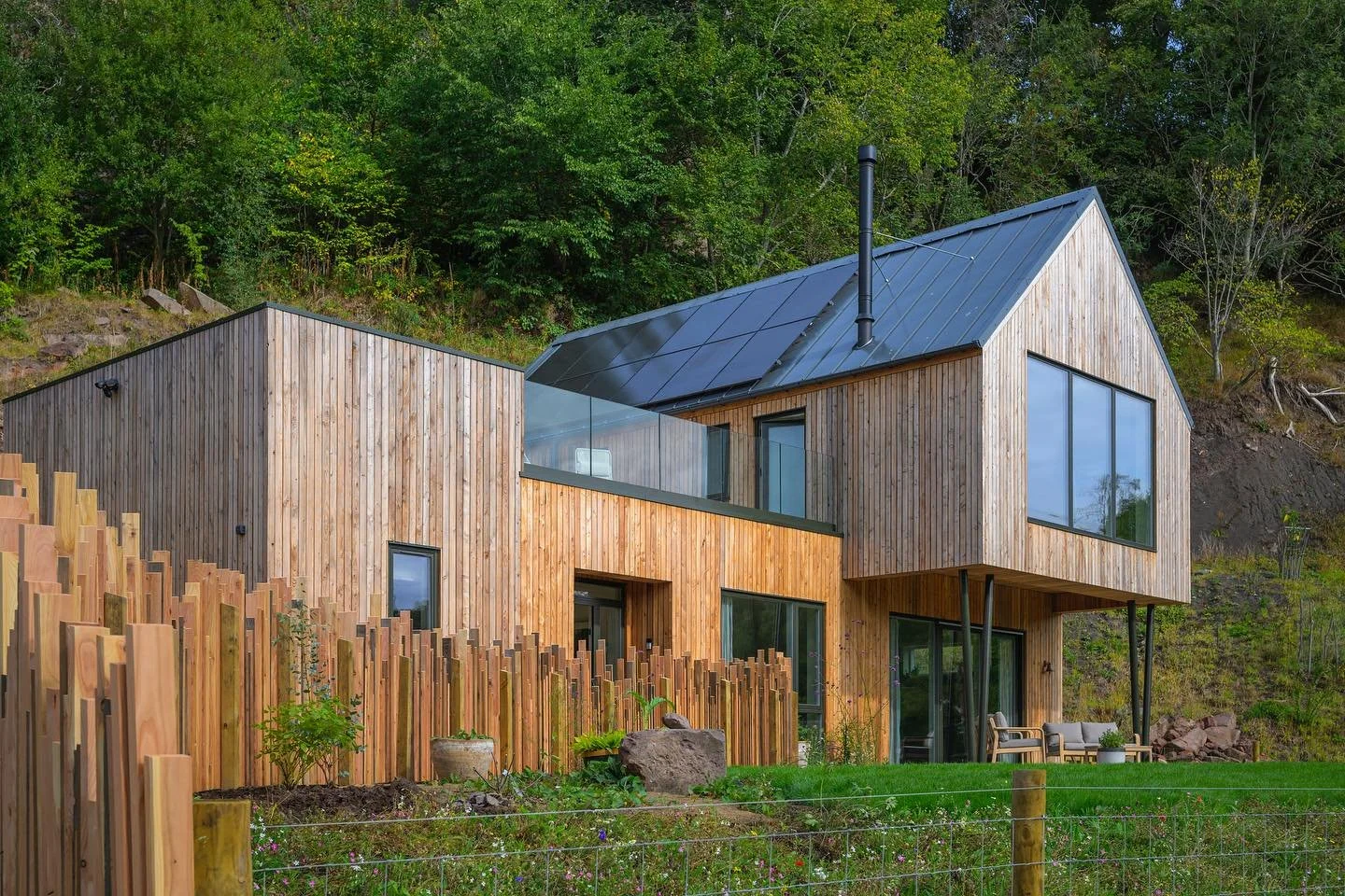 Modern two-story house with a wooden exterior, large windows, a black chimney, and a metal roof, situated in a green natural landscape.