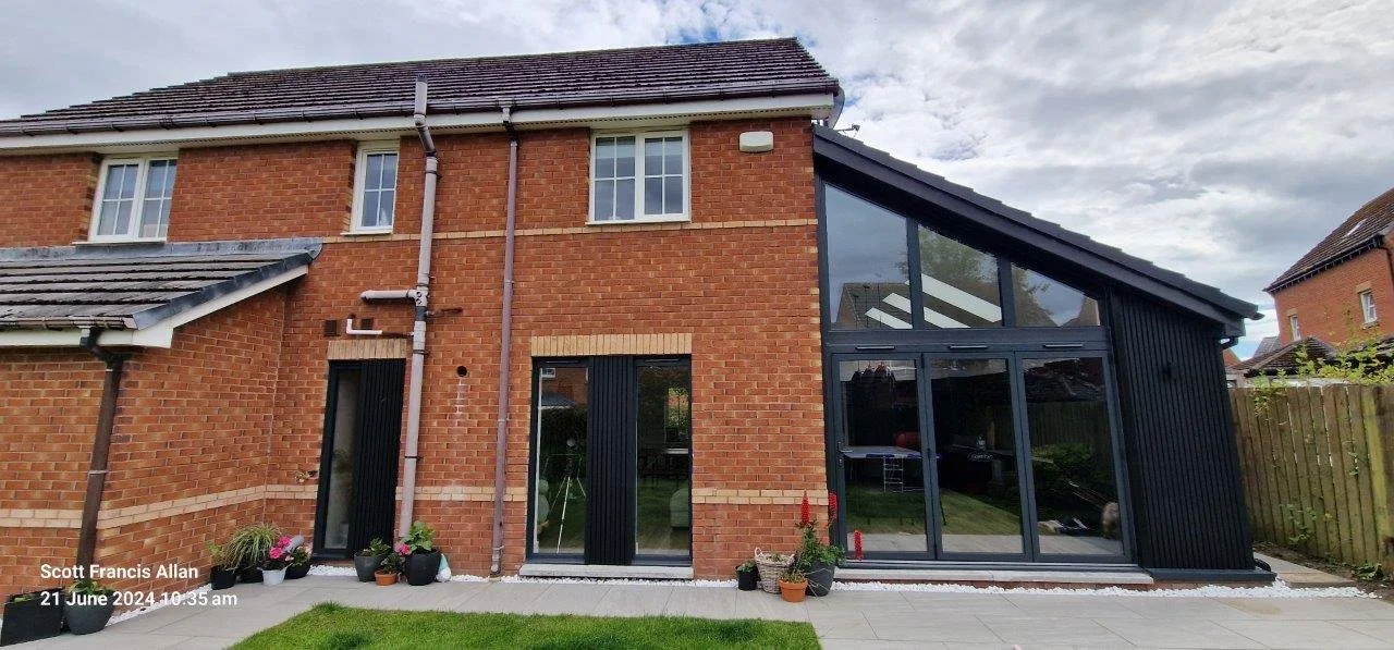Back view of a modern two-story brick house with a black extension featuring large glass windows and doors, and potted plants on the patio. Overcast sky.