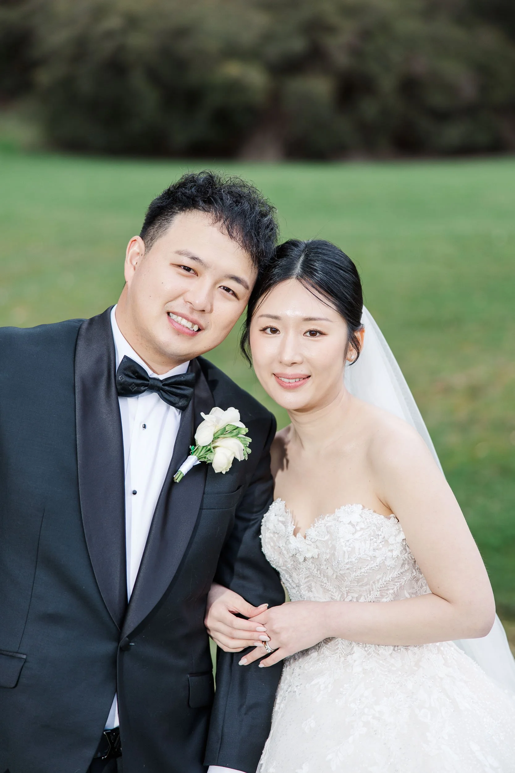 Melbourne Asian Wedding photography of a smiling bride and groom in formalwear, posed outdoors with soft light and a green backdrop.