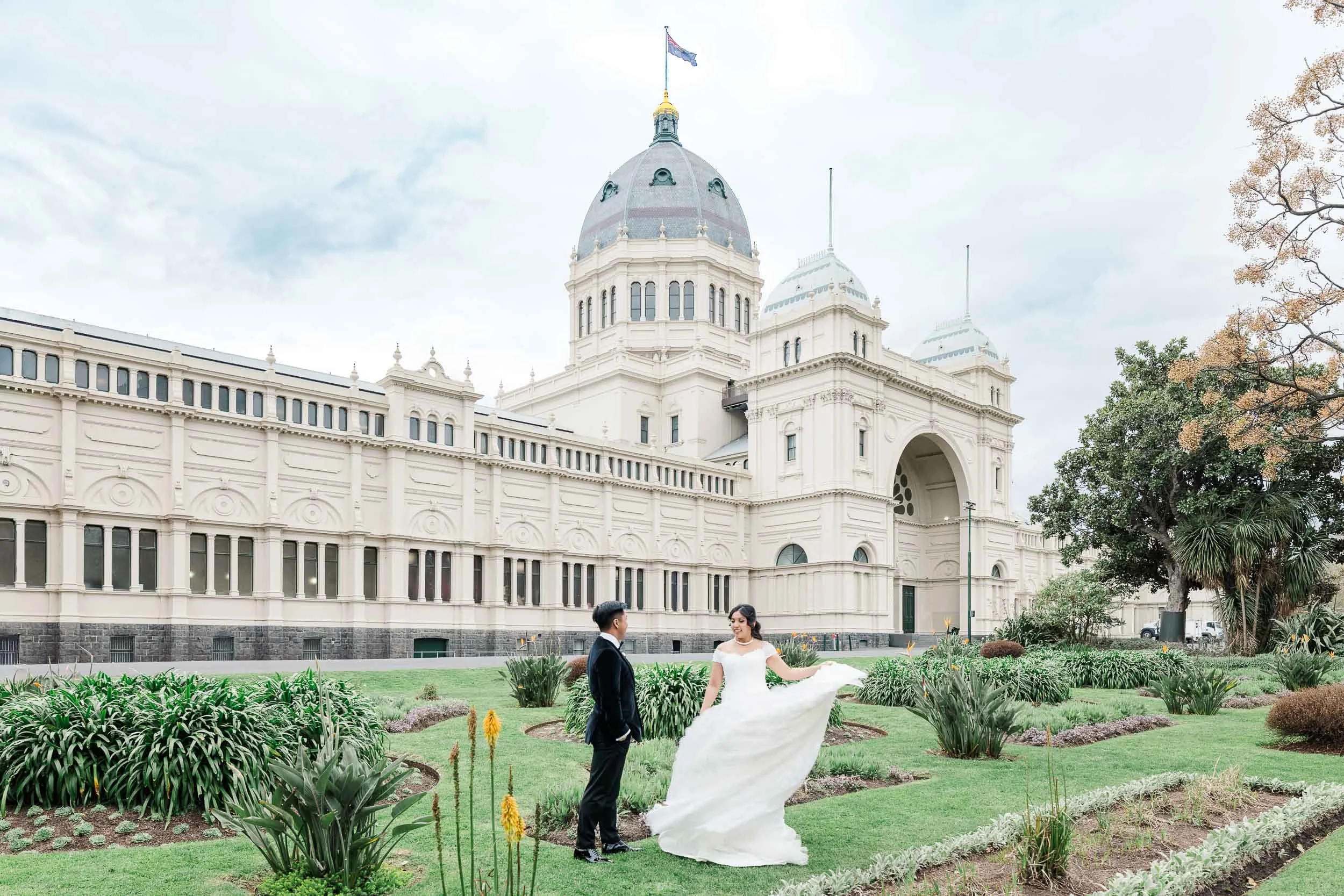 Melbourne wedding photography of a bride and groom outside the Royal Exhibition Building, framed by elegant heritage architecture and gardens.