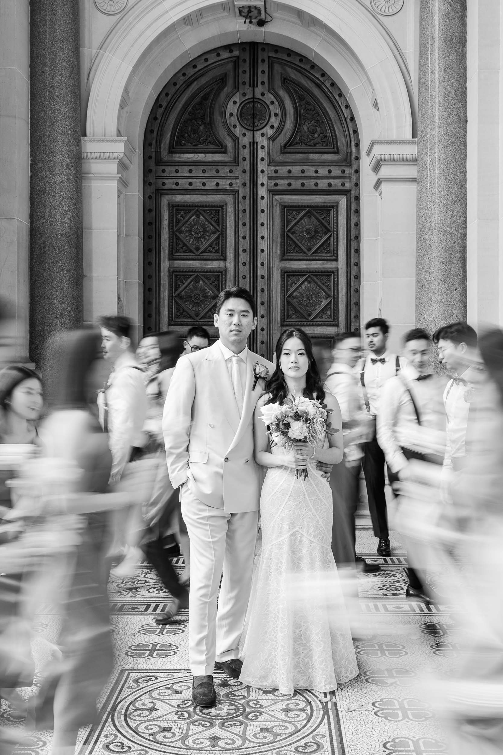 Black and white Melbourne wedding photography of Audrey and Fenly standing still in a grand doorway as guests blur past around them.